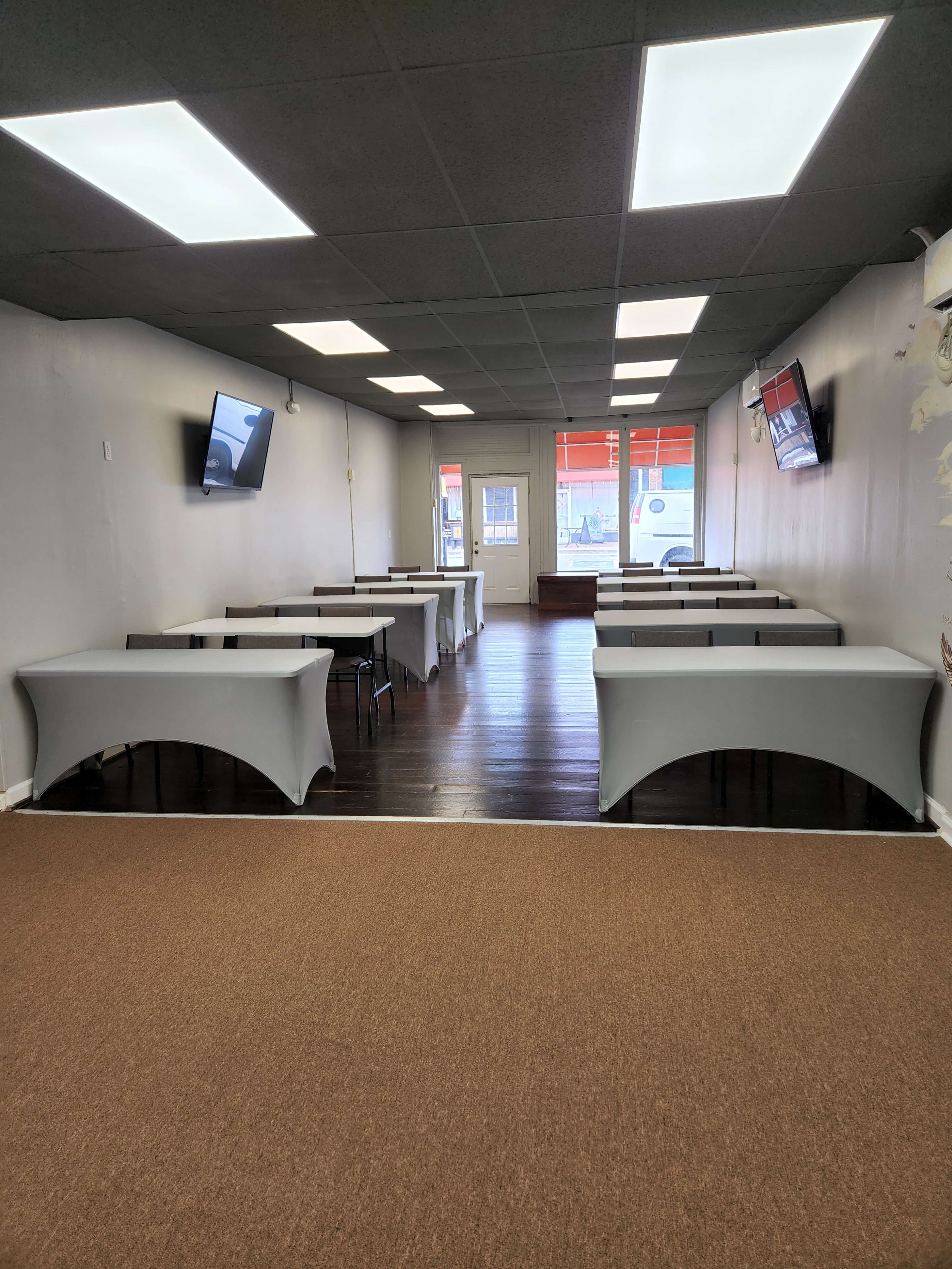 An interior of a room with several tables covered in white tablecloths and two wall-mounted screens, leading to a door at the far end.