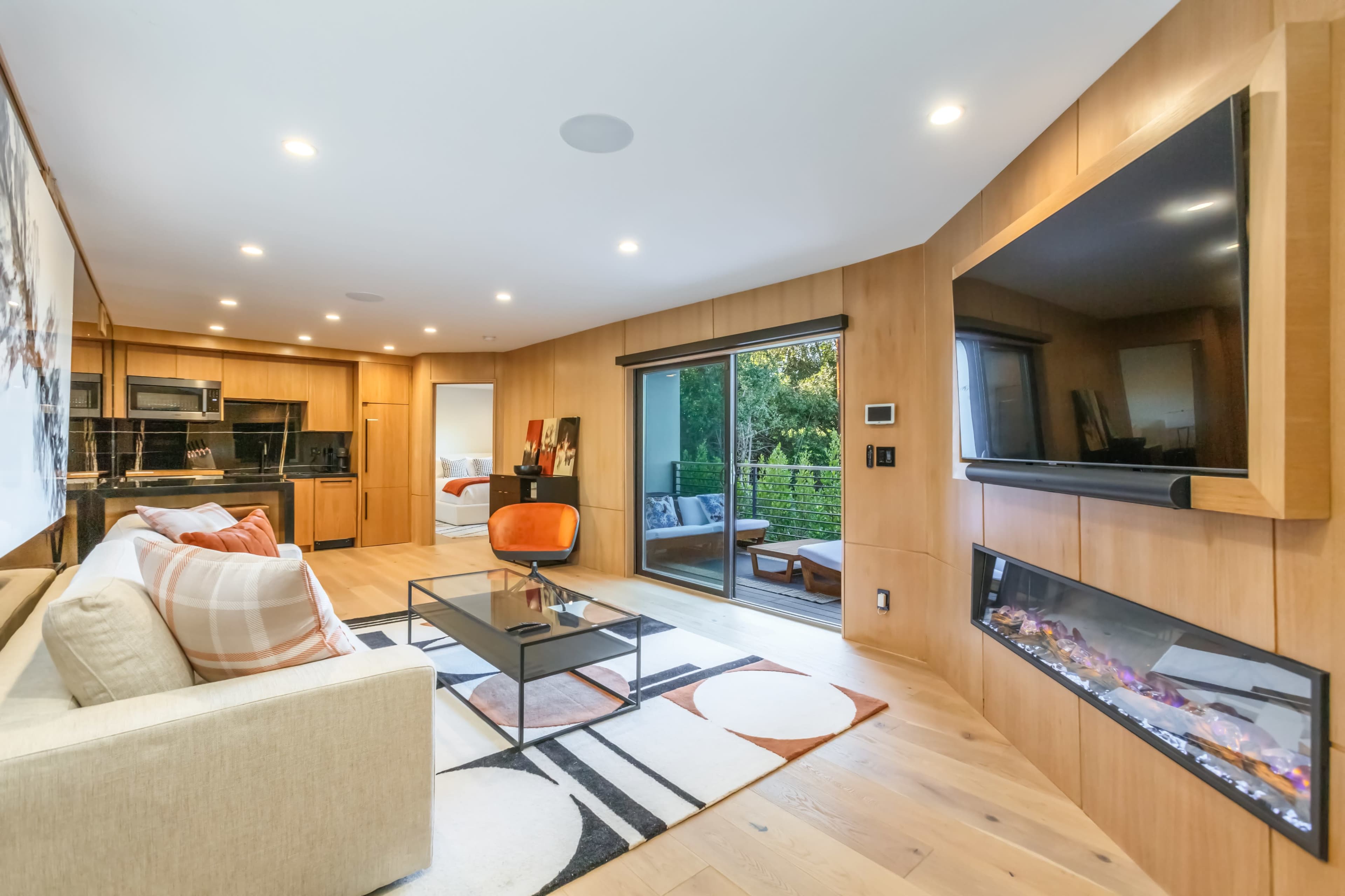 A modern living room features a large window, a flat-screen TV mounted on the wall, and a cozy seating area with a beige couch and a coffee table.