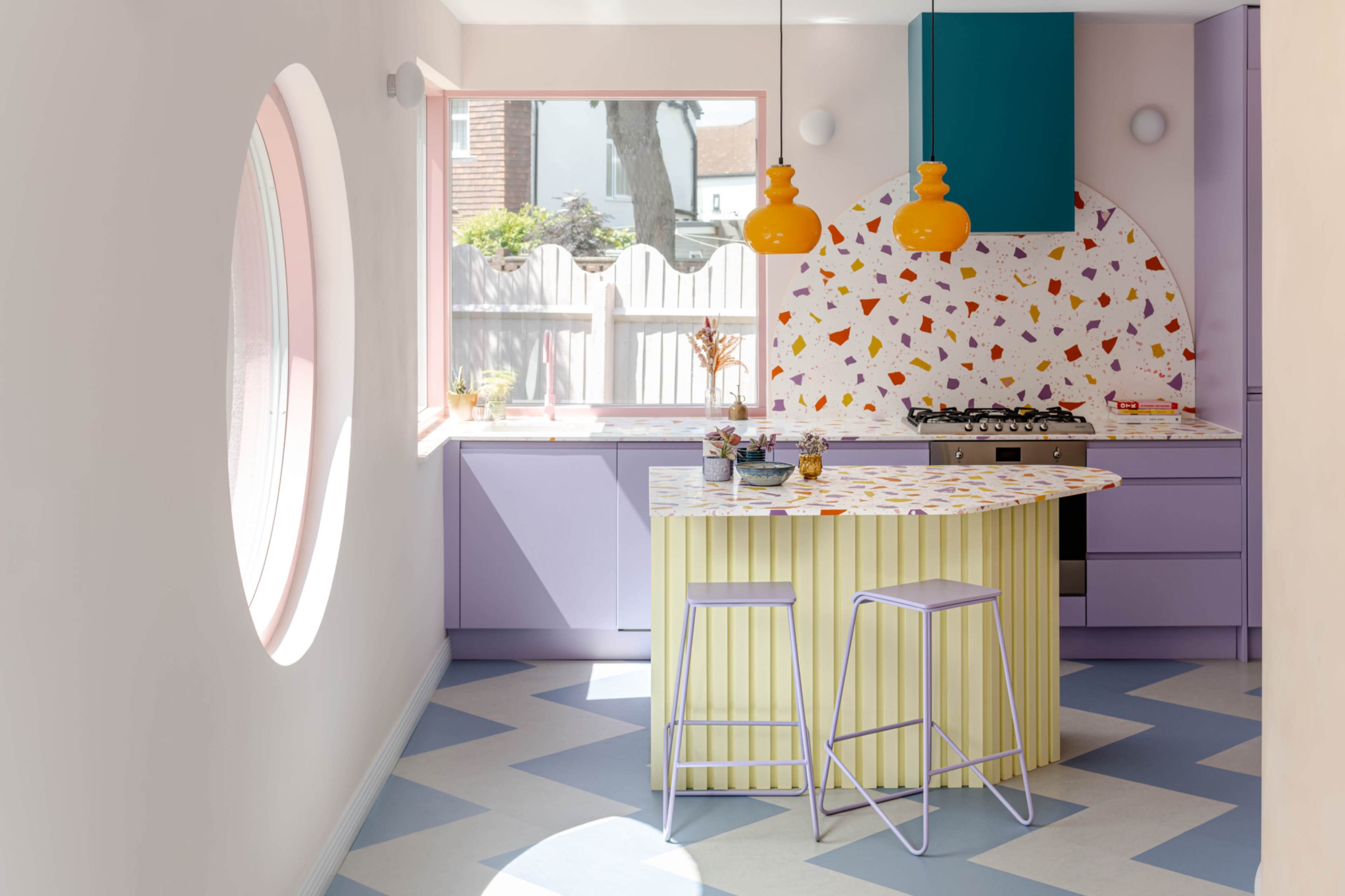 The image showcases a brightly designed kitchen featuring a curved island with a yellow and white striped base, purple cabinetry, and colorful decorative elements on the backsplash.