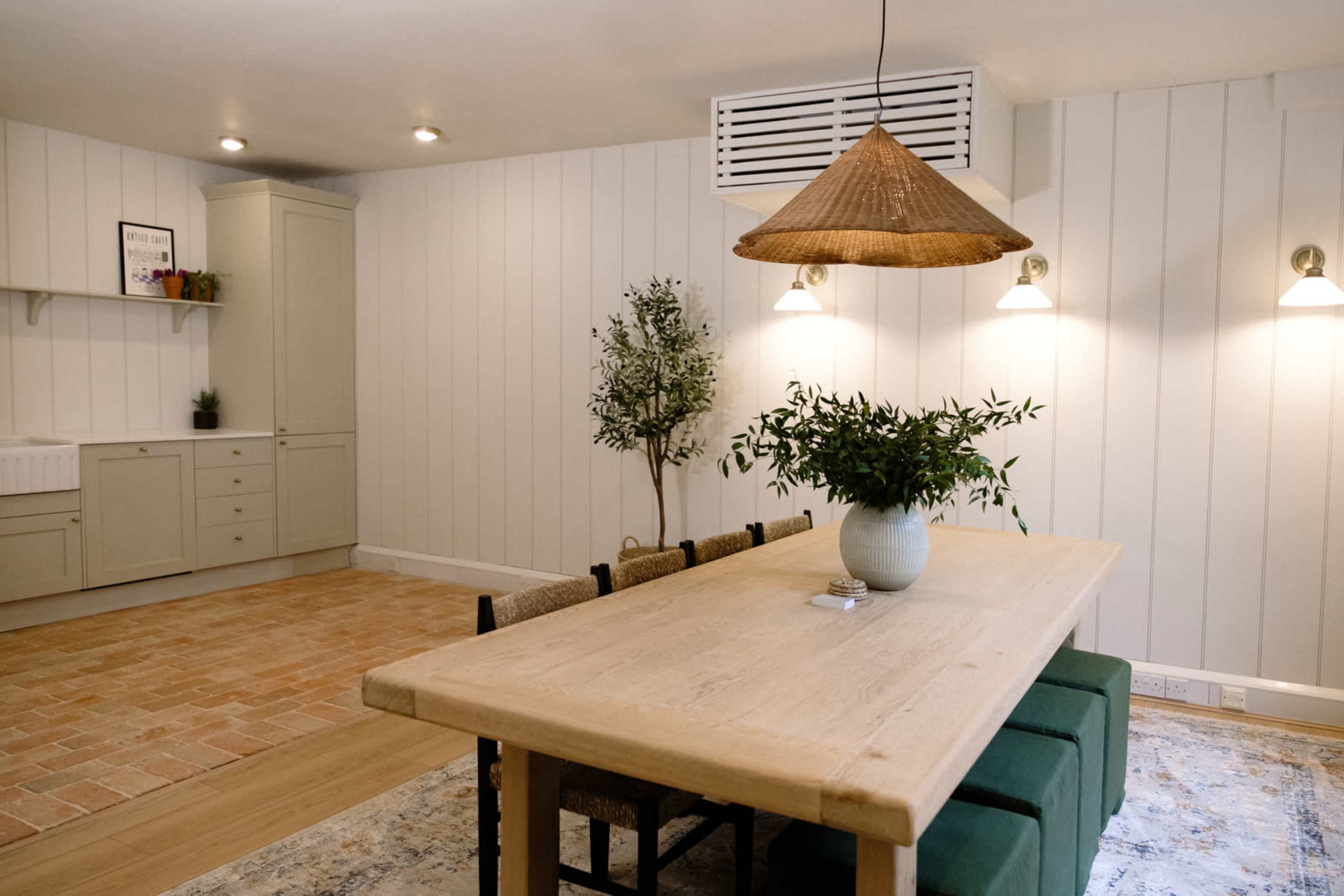 A dining area features a wooden table with chairs, a potted plant, and wall-mounted lights, set against light-colored paneled walls and a brick floor.