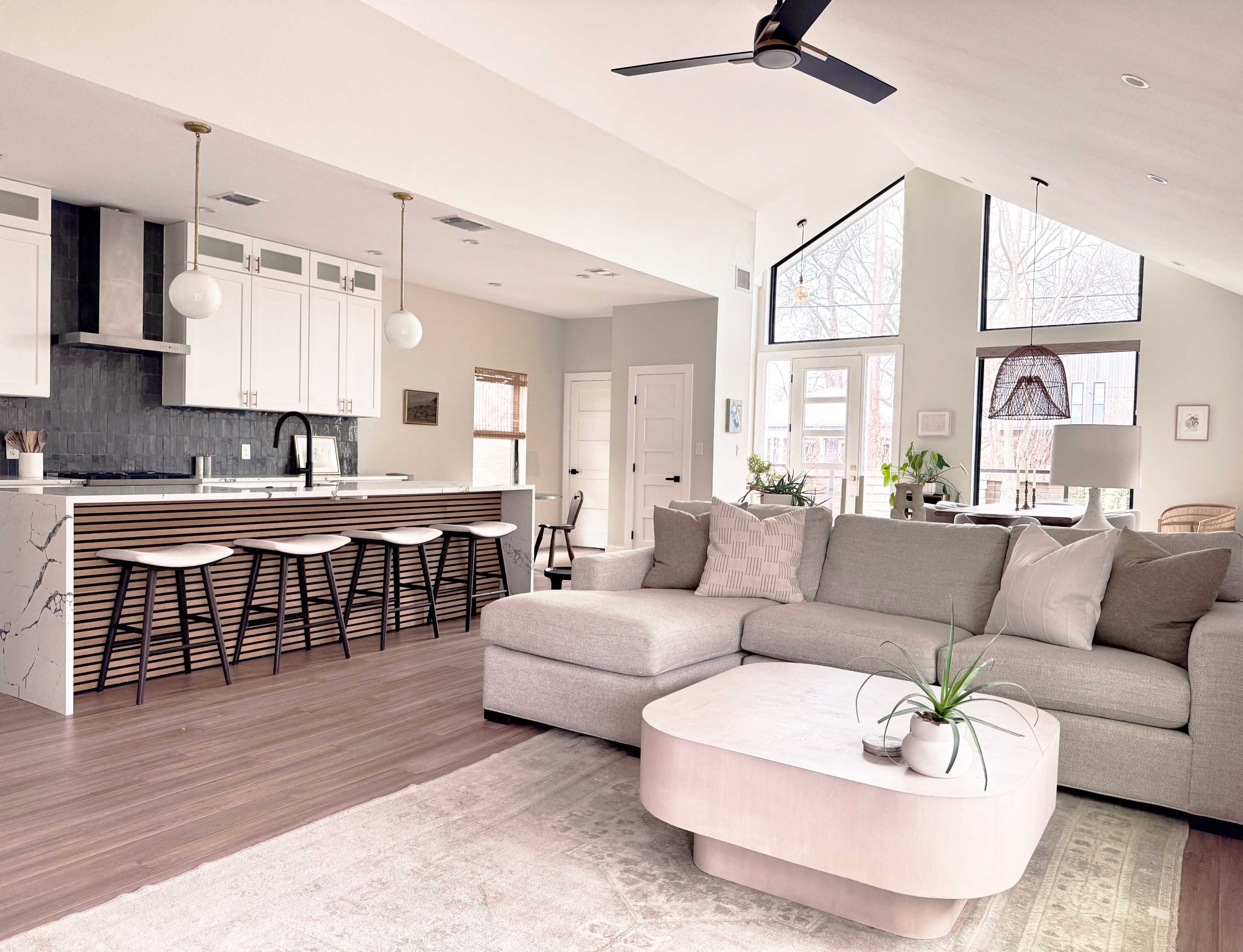 The image shows a modern living room and kitchen area featuring a large gray sectional sofa, a round coffee table, and a white kitchen island with bar stools.