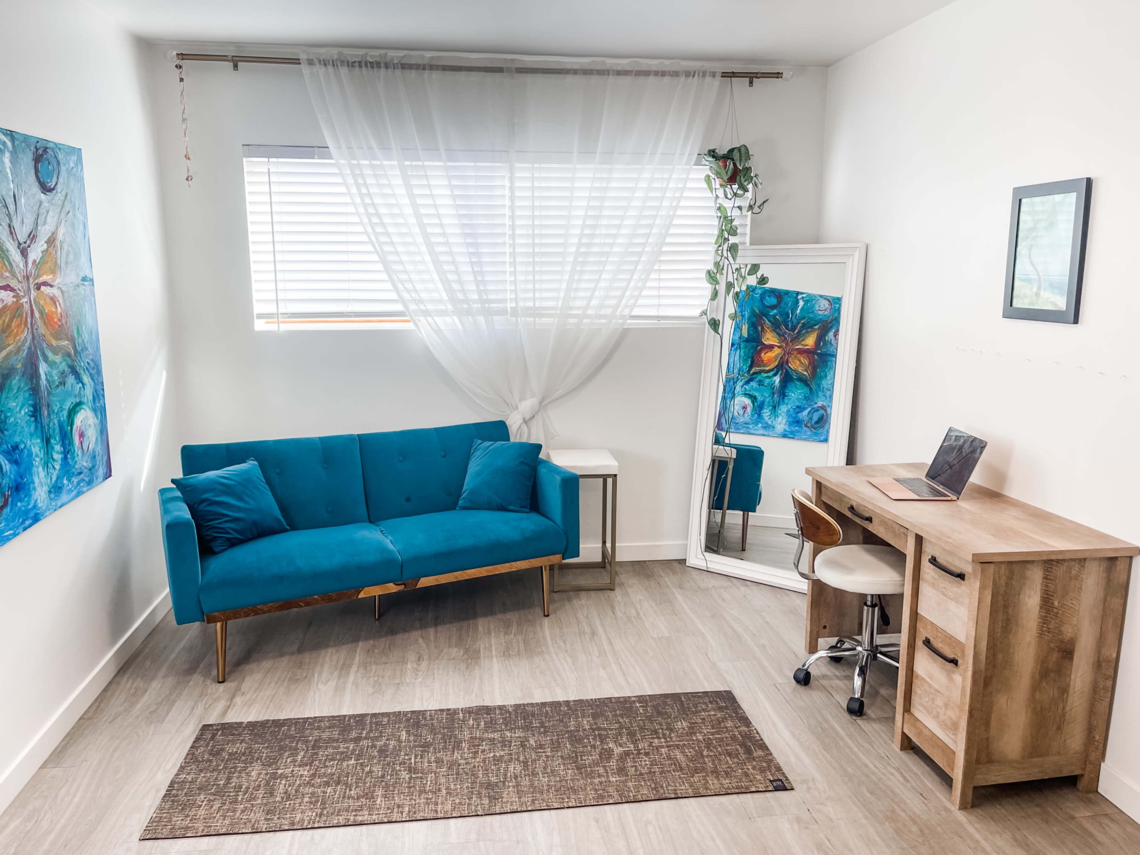 A modern room with a turquoise couch, a wooden desk and chair, a mirror, and a colorful painting on the wall.