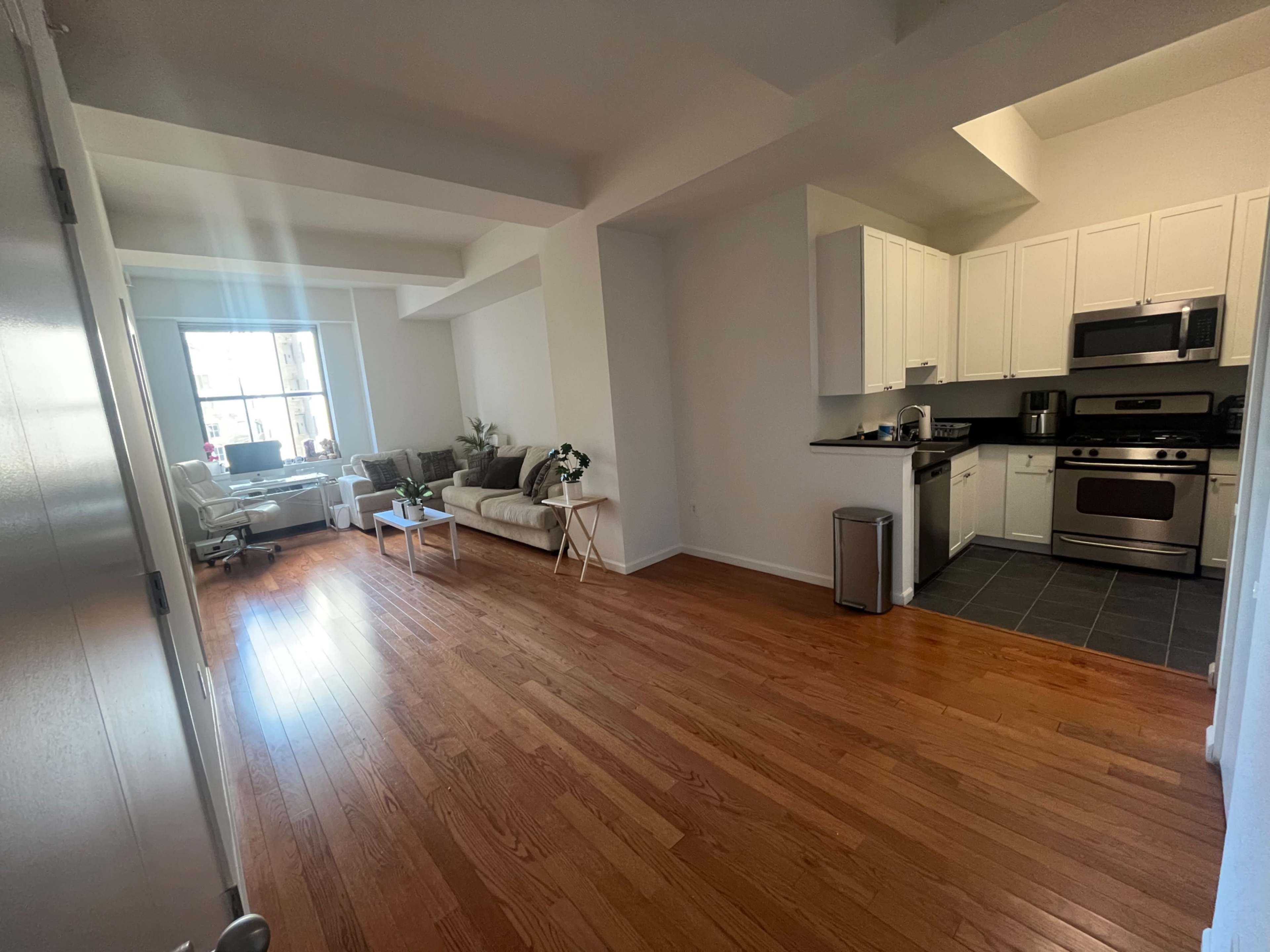The image shows a modern, open living space with wooden floors, a kitchen area, and a seating area featuring a sofa and a small table.