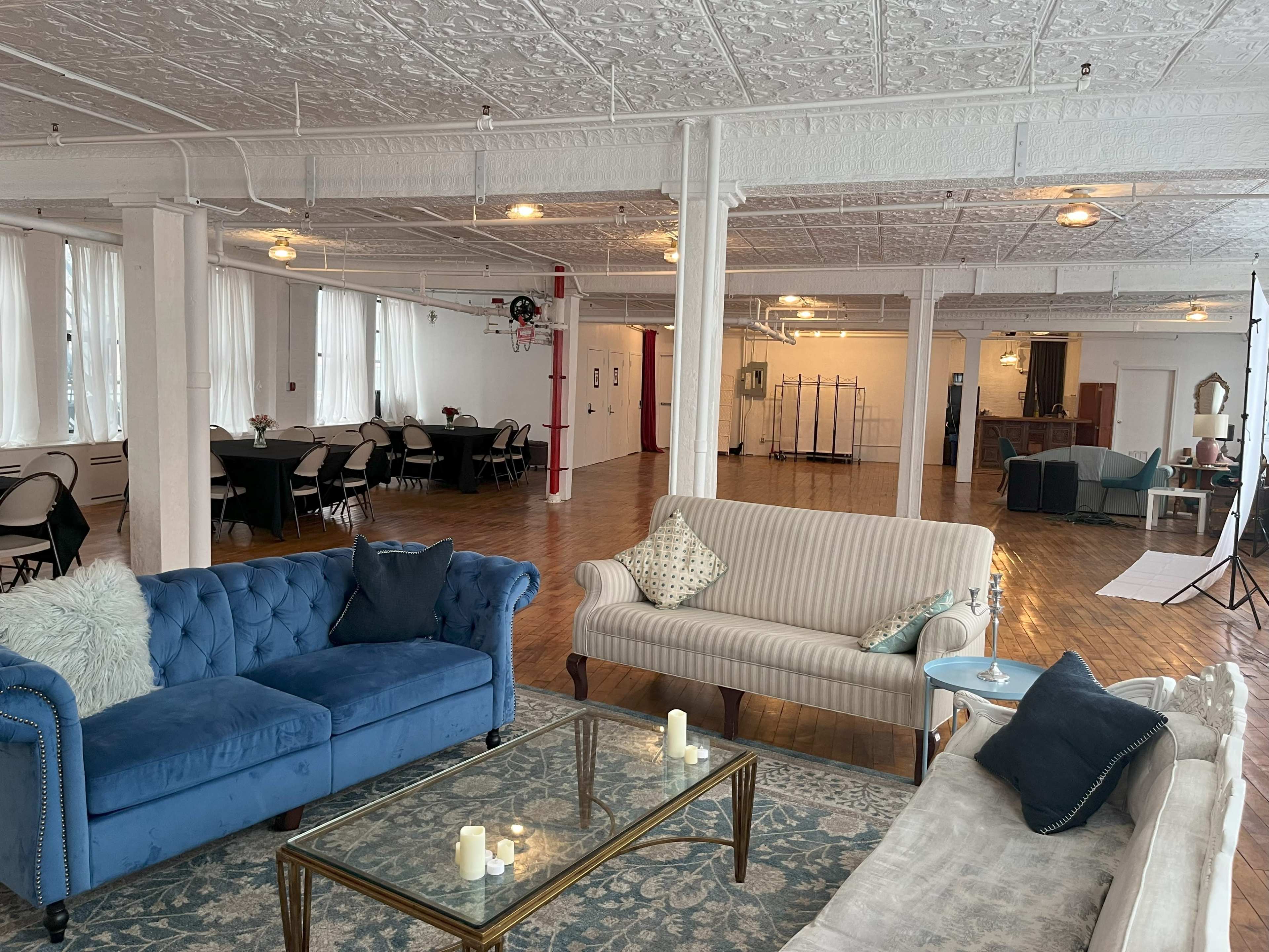 A spacious, well-lit room with a seating area featuring blue and beige couches, a glass coffee table, and several tables set up in the background for an event.