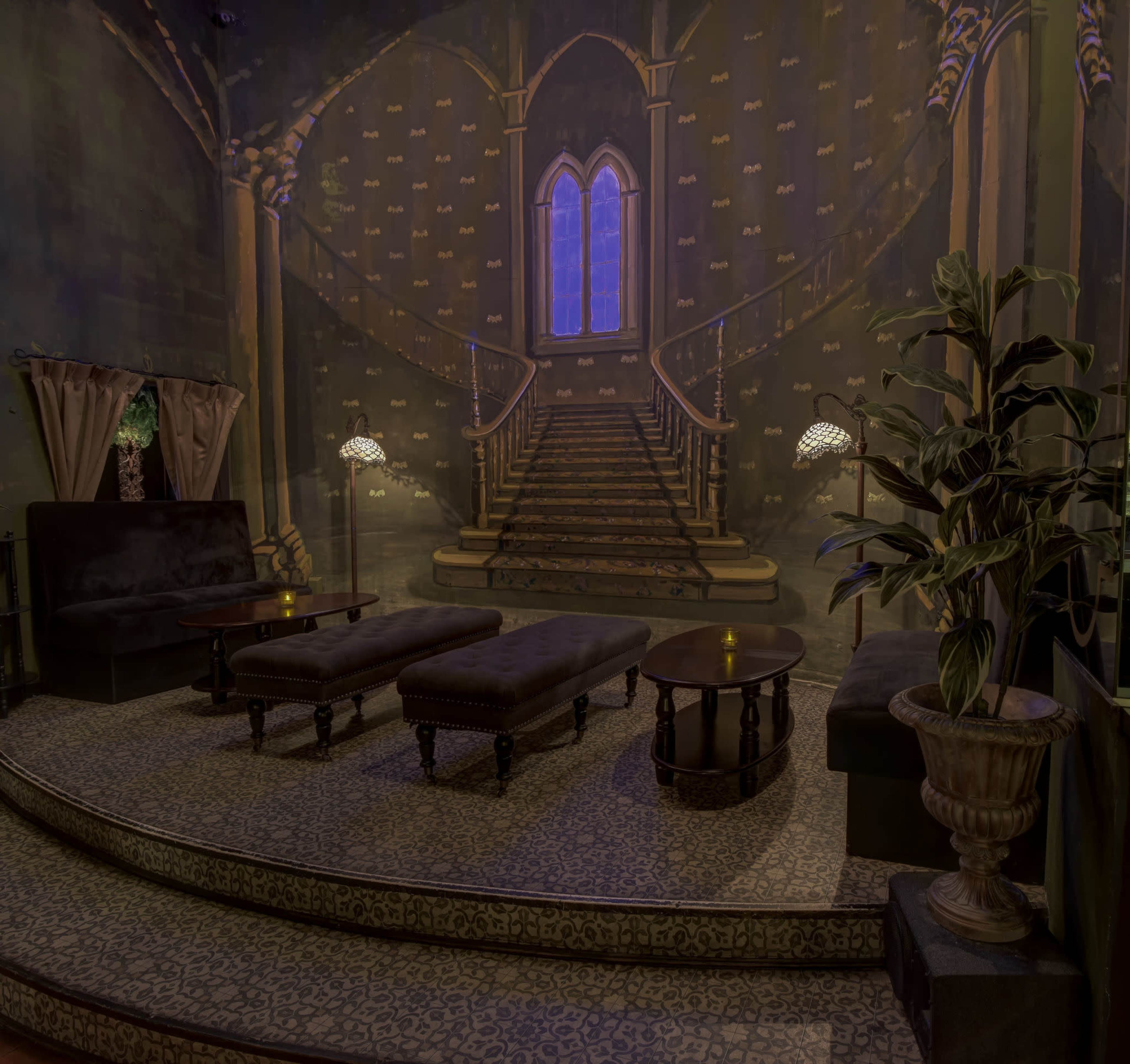 The image shows a dimly lit room with a mural depicting a grand staircase and two lamps beside low furniture, including benches and a table, surrounded by decorative plants.