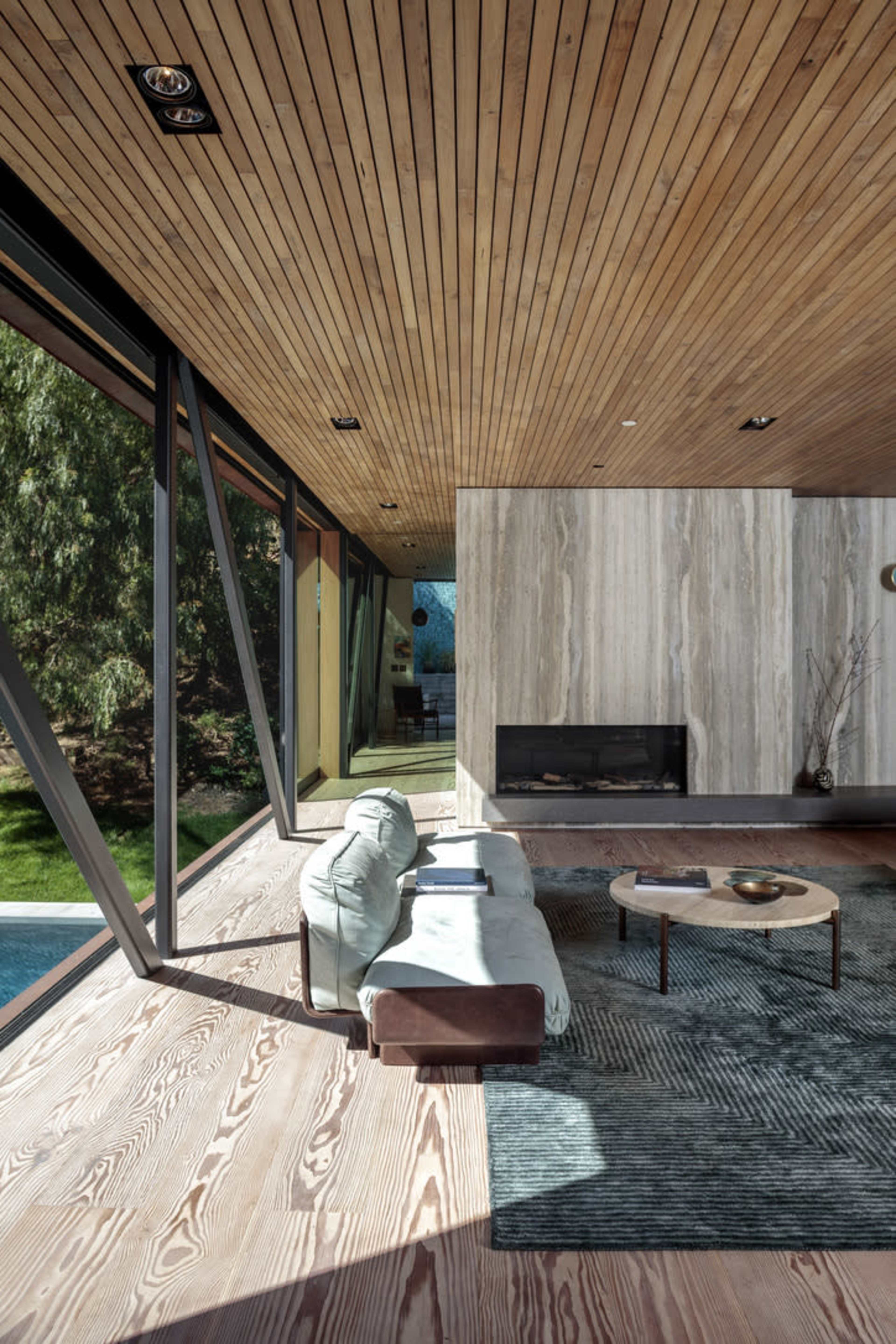 The interior of a modern living room features wooden ceilings, large windows, a minimalist sofa, and a stone fireplace.