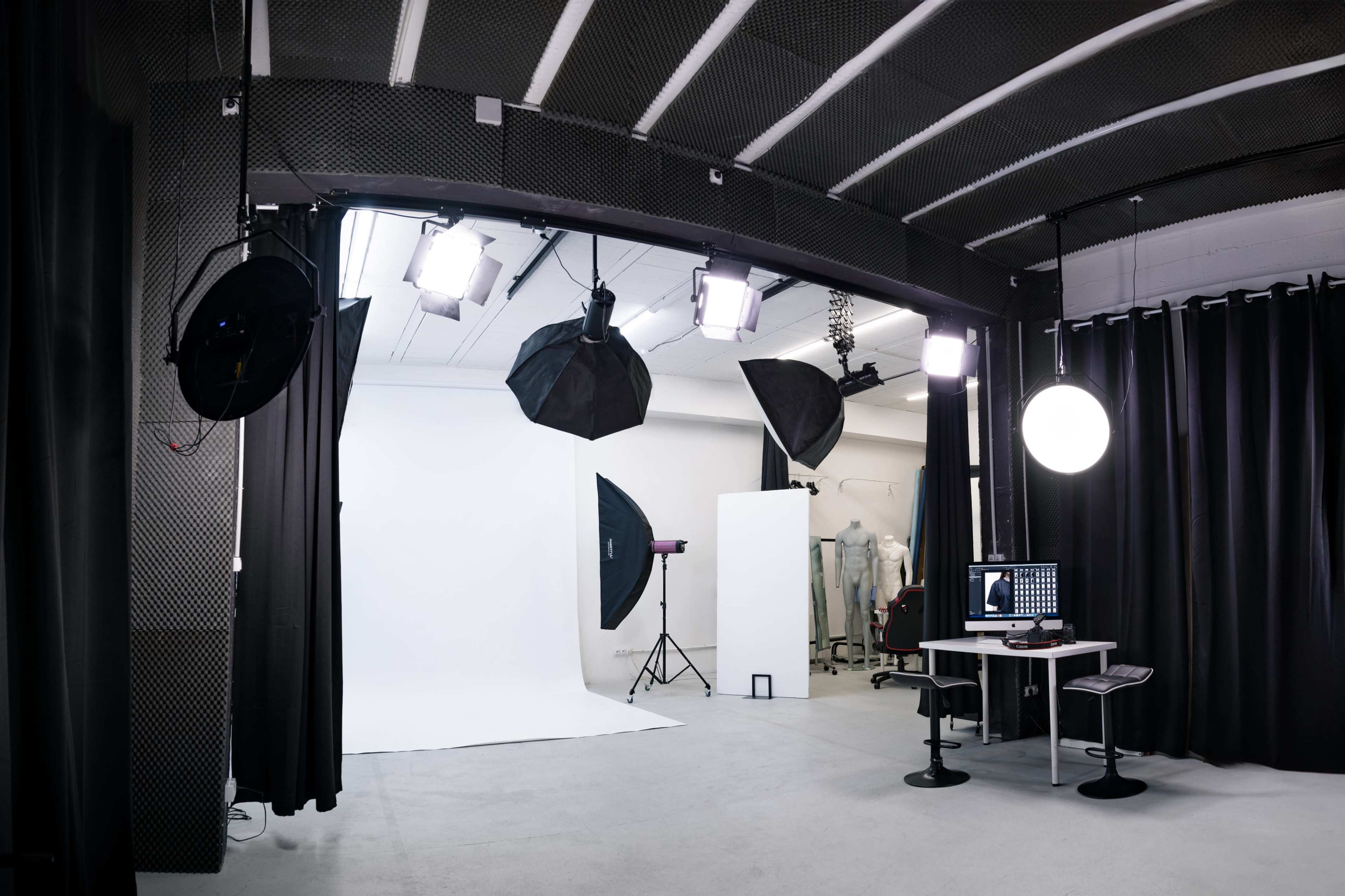 A photography studio features a white backdrop, multiple light sources, a computer setup, and dark curtains.