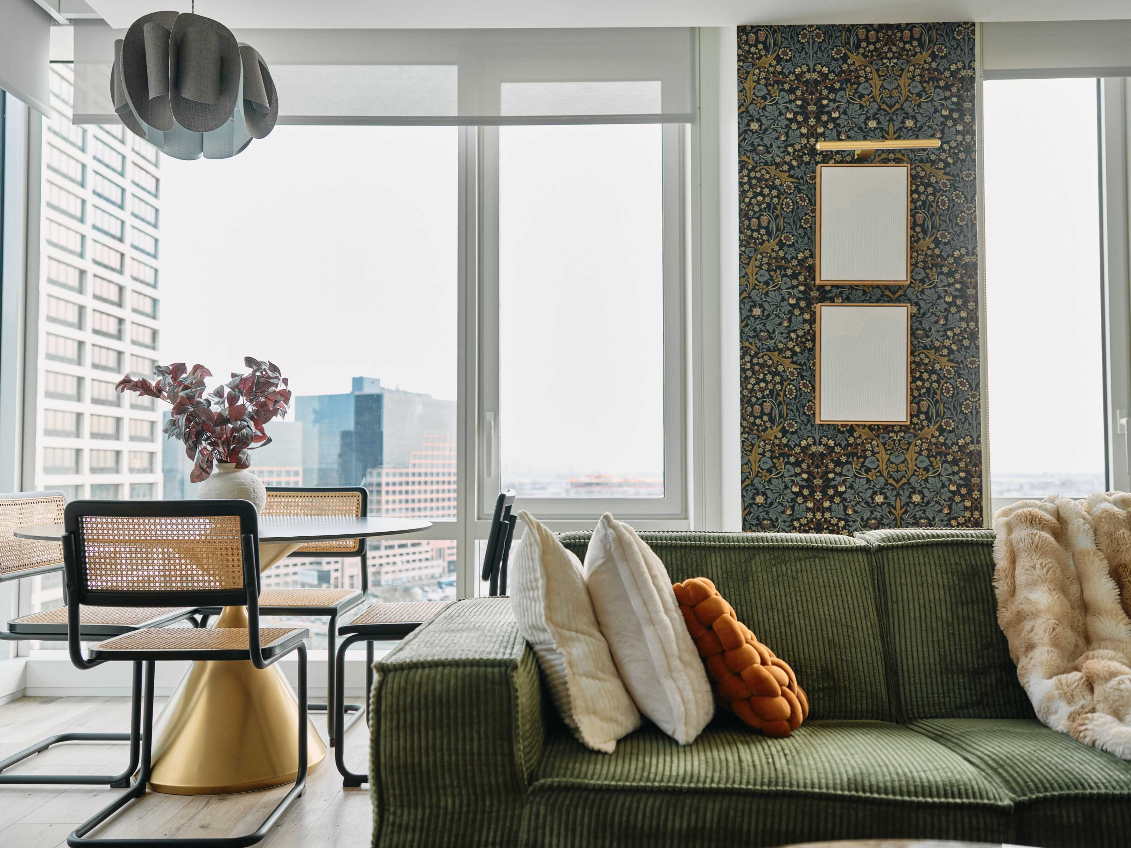 A modern living room features a green sofa with decorative pillows, a dining area with a round table and chairs, and large windows showcasing a city skyline.