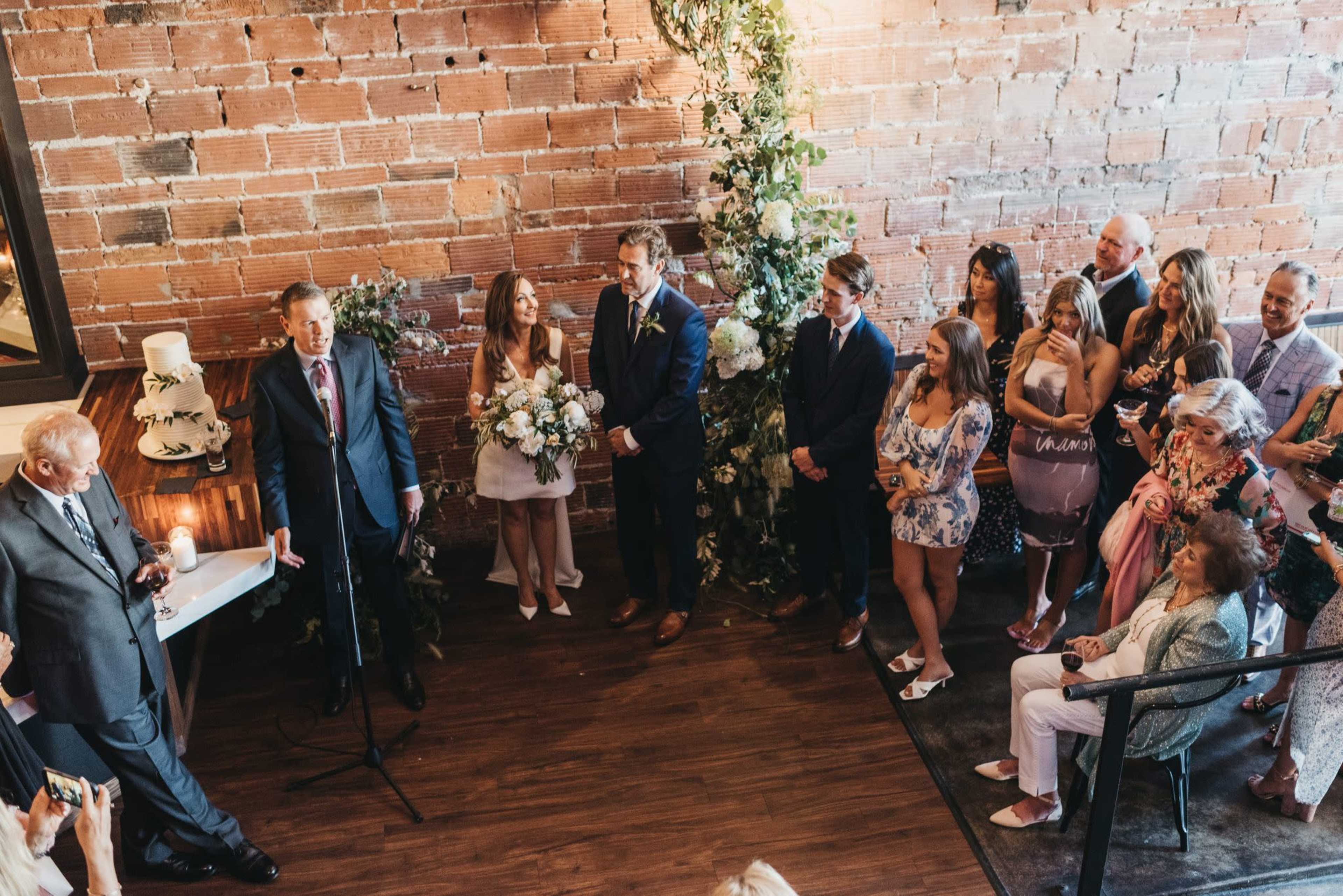 A couple stands at the front of a brick-walled venue, surrounded by guests as they participate in a wedding ceremony.