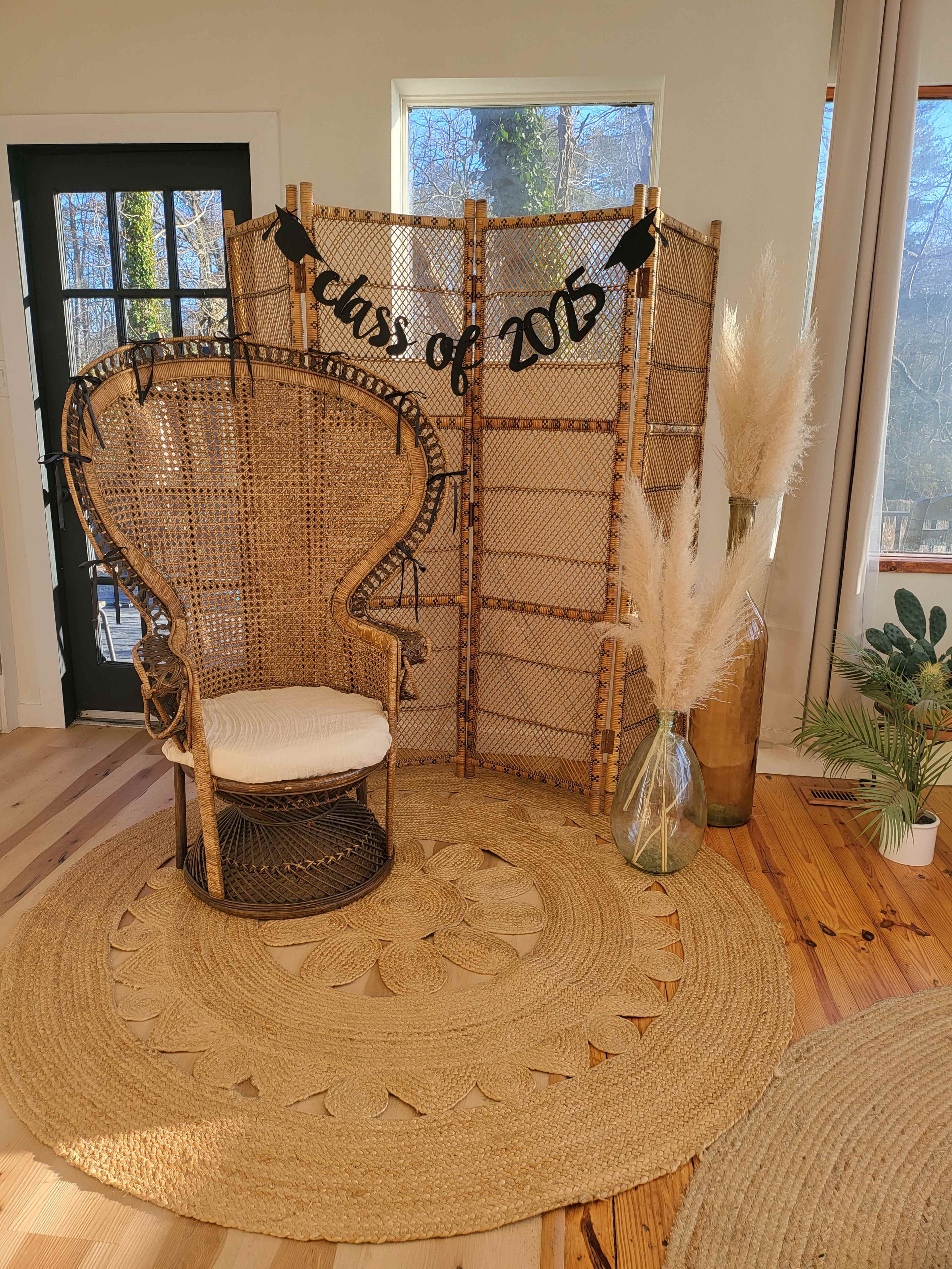 A wicker chair is positioned in front of a decorative screen that reads "Class of 2025," with a vase of pampas grass nearby on a round woven rug.