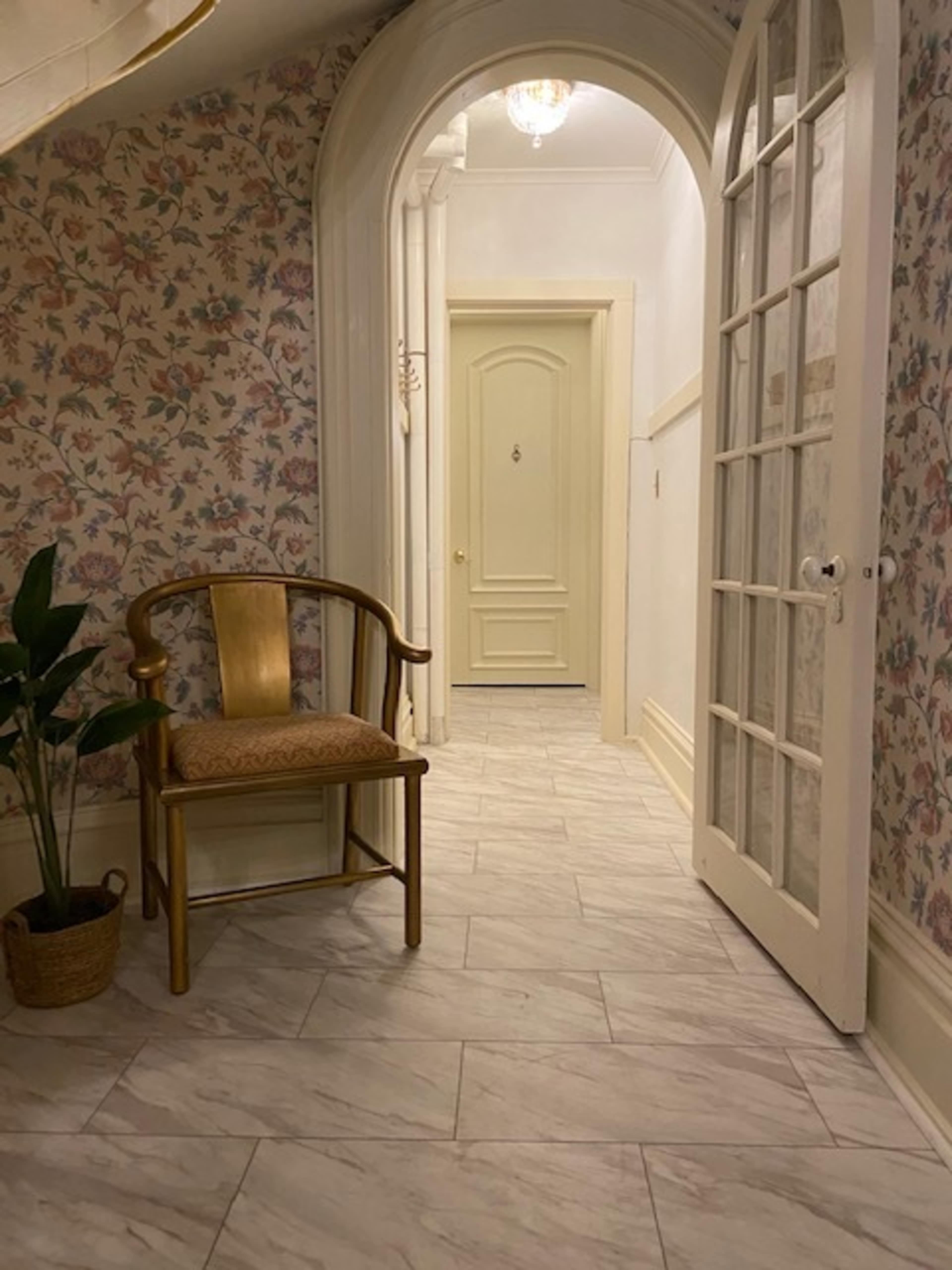 A hallway with floral wallpaper, a wooden chair near a potted plant, and a door at the far end.