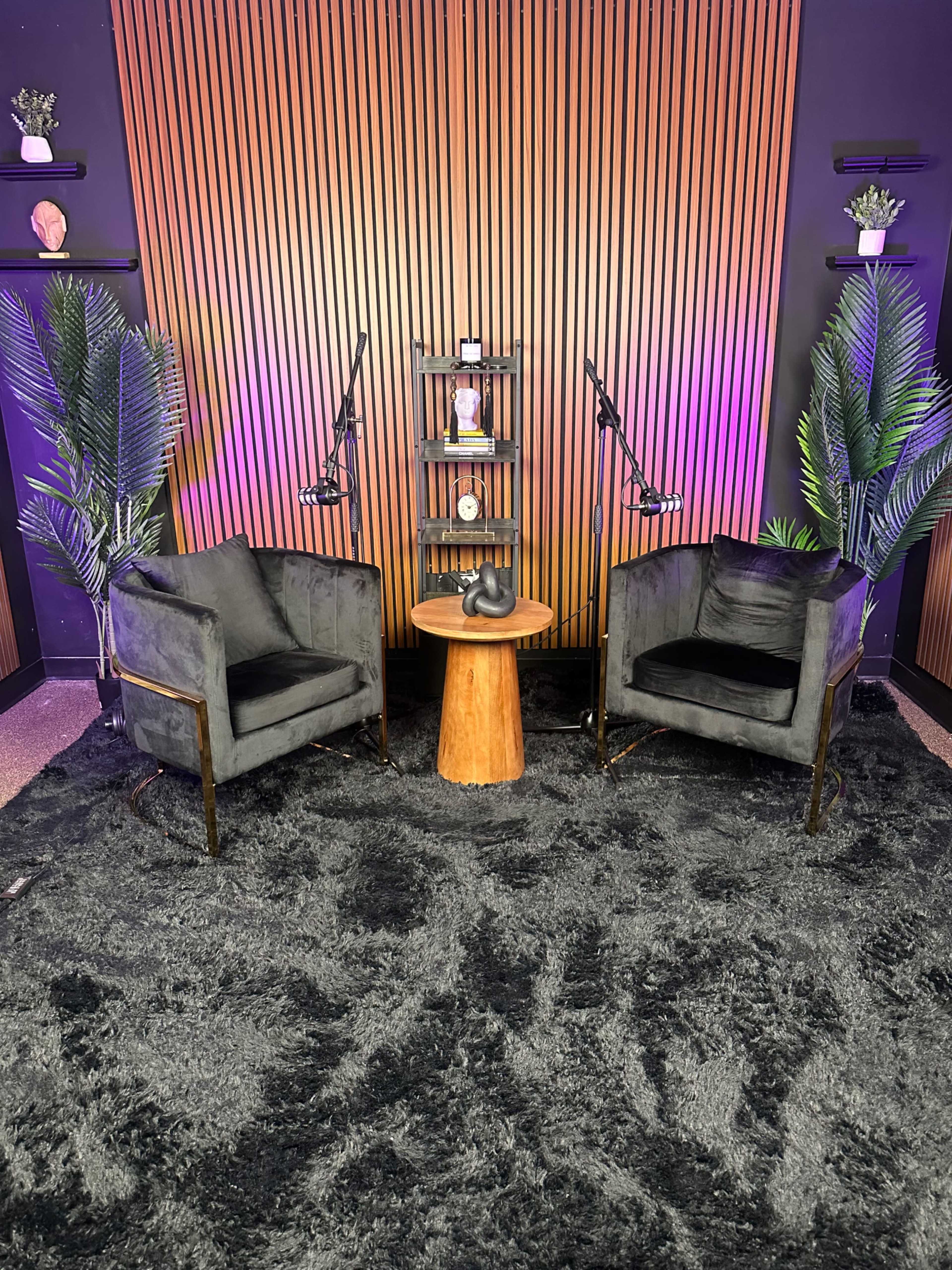 The image depicts a cozy seating area with two black chairs facing a wooden side table, set against a backdrop of vertical wooden slats and soft purple lighting.
