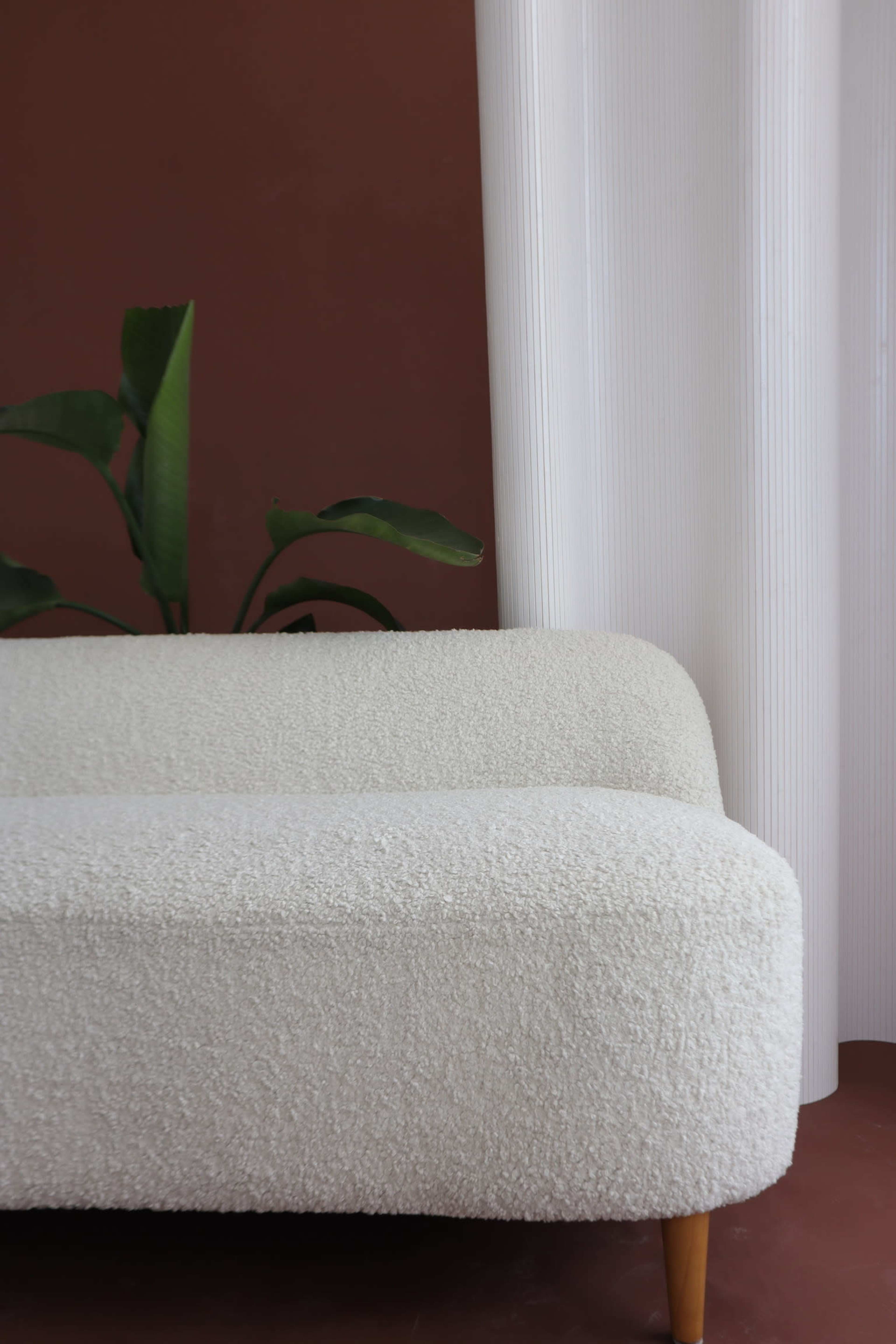 A white, textured sofa with rounded edges sits against a brown wall, next to a green plant and sheer white curtains.