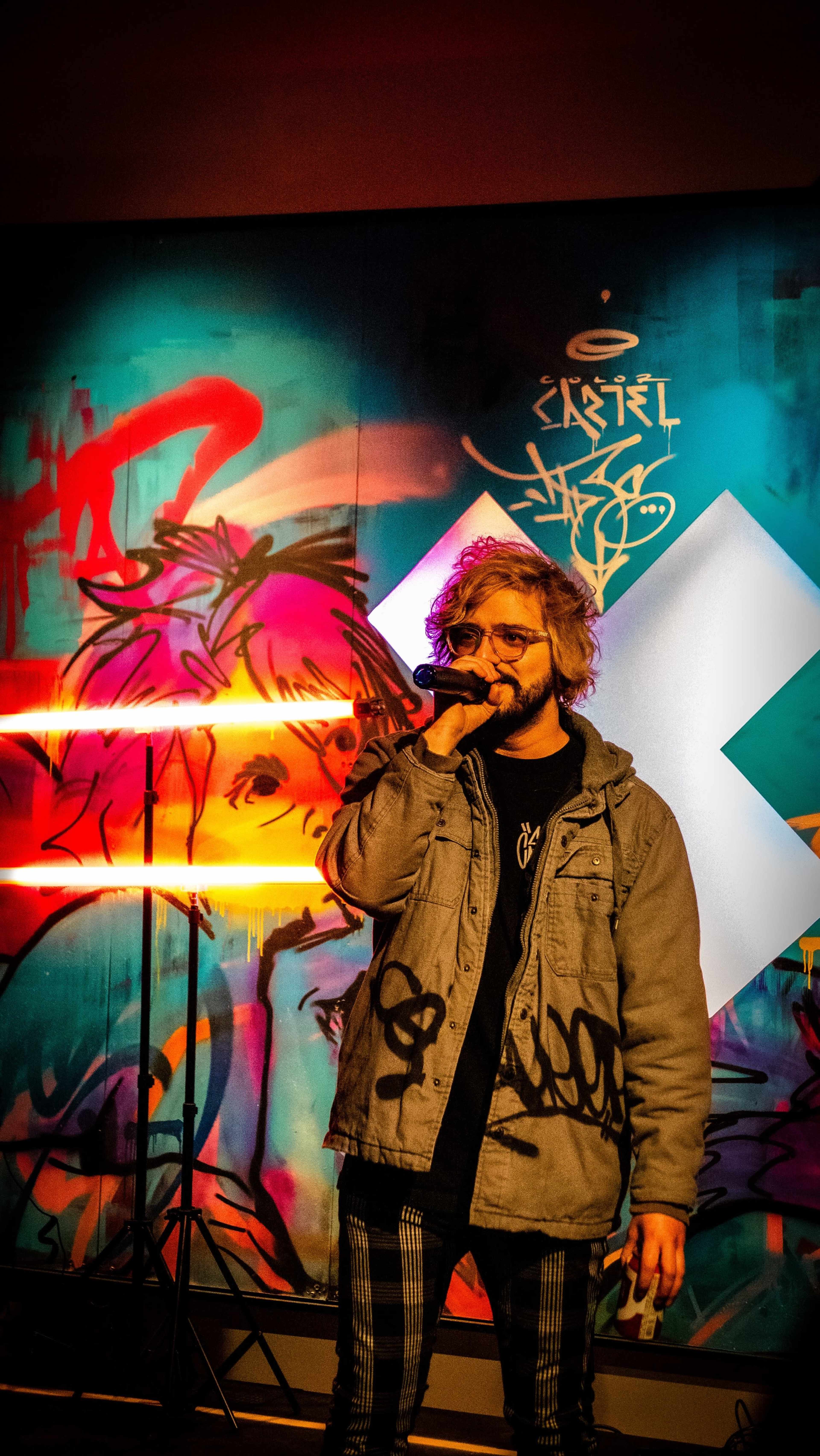 A person holds a microphone while performing in front of a colorful graffiti backdrop.