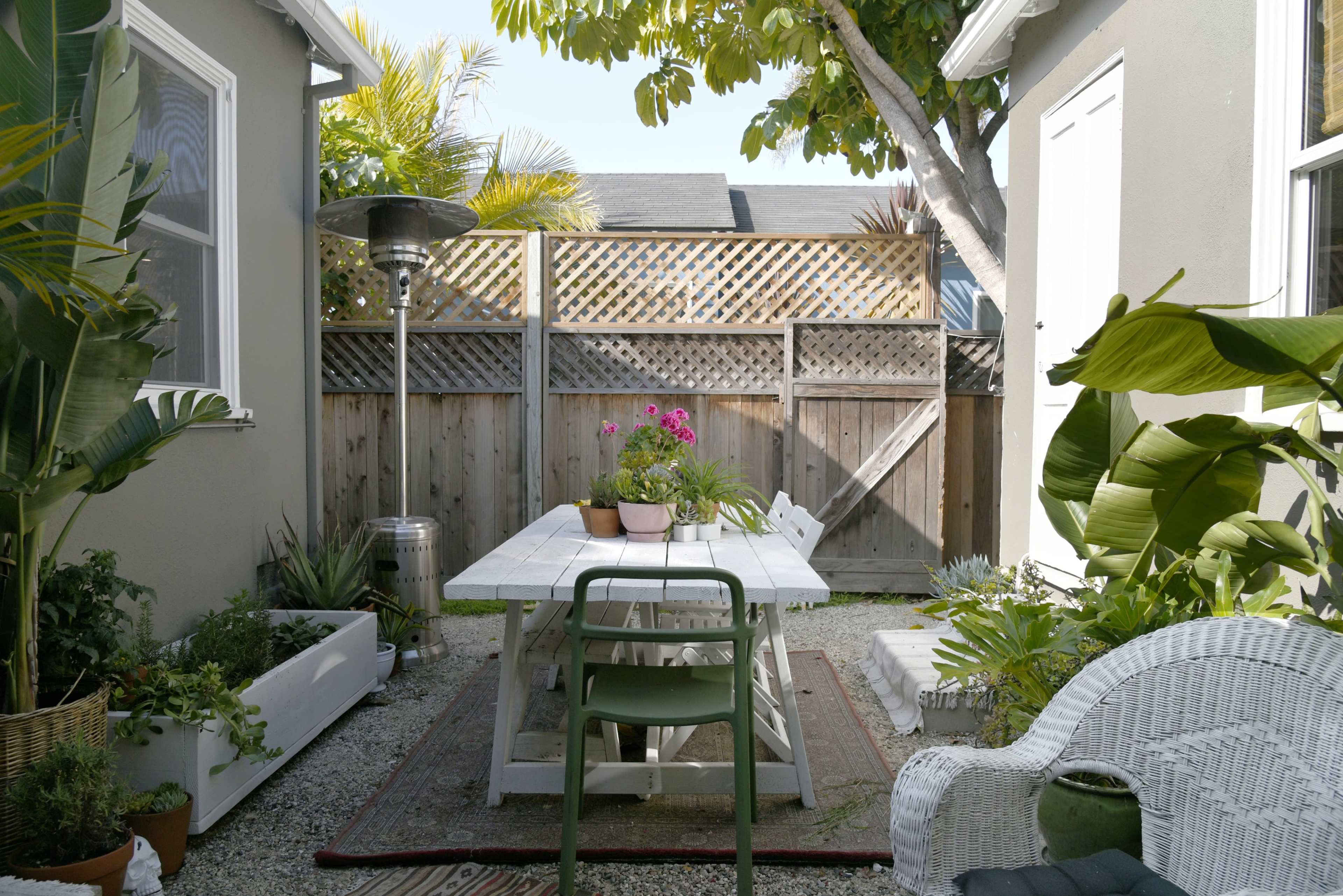 A small outdoor patio features a white table surrounded by chairs, with potted plants and a heater, enclosed by wooden fencing.