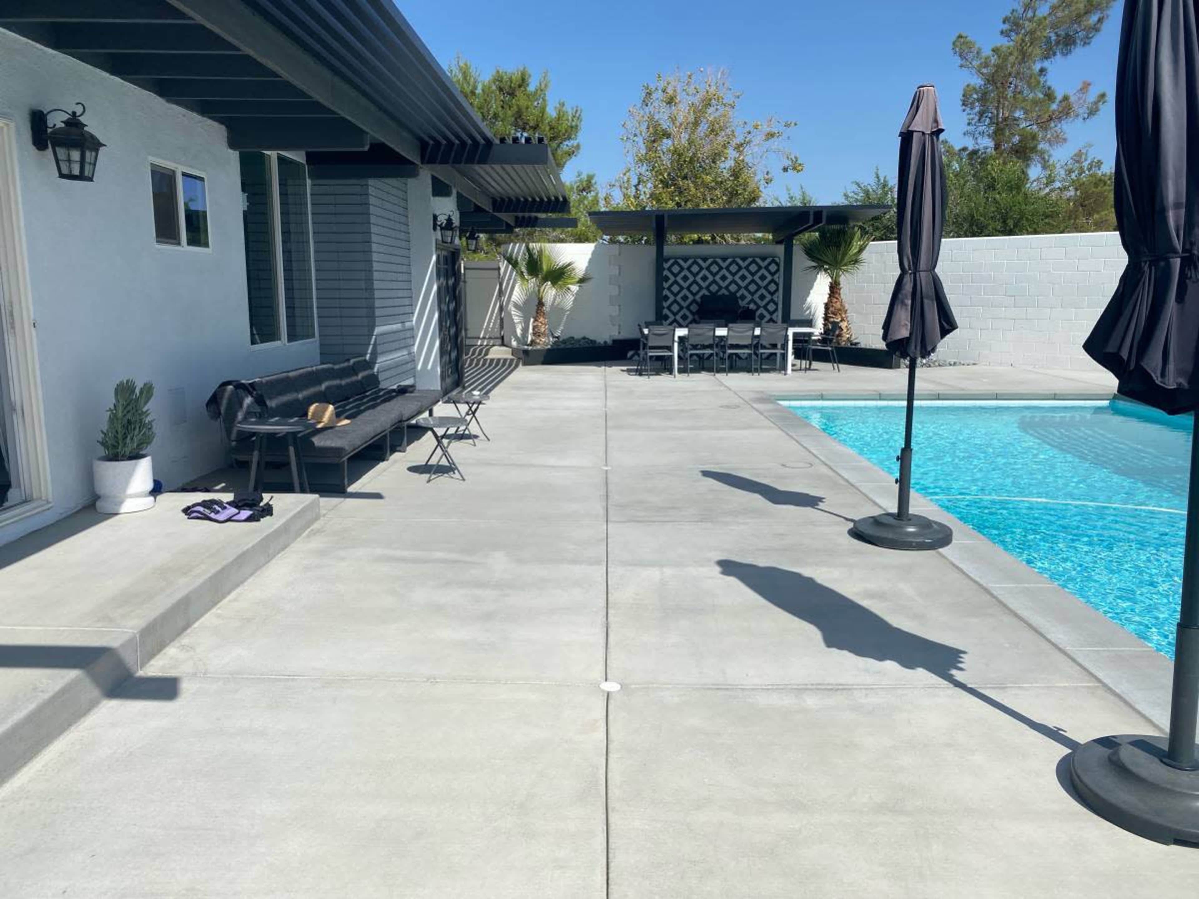 A modern backyard featuring a swimming pool, lounge chairs, and patio umbrellas against a clear blue sky.