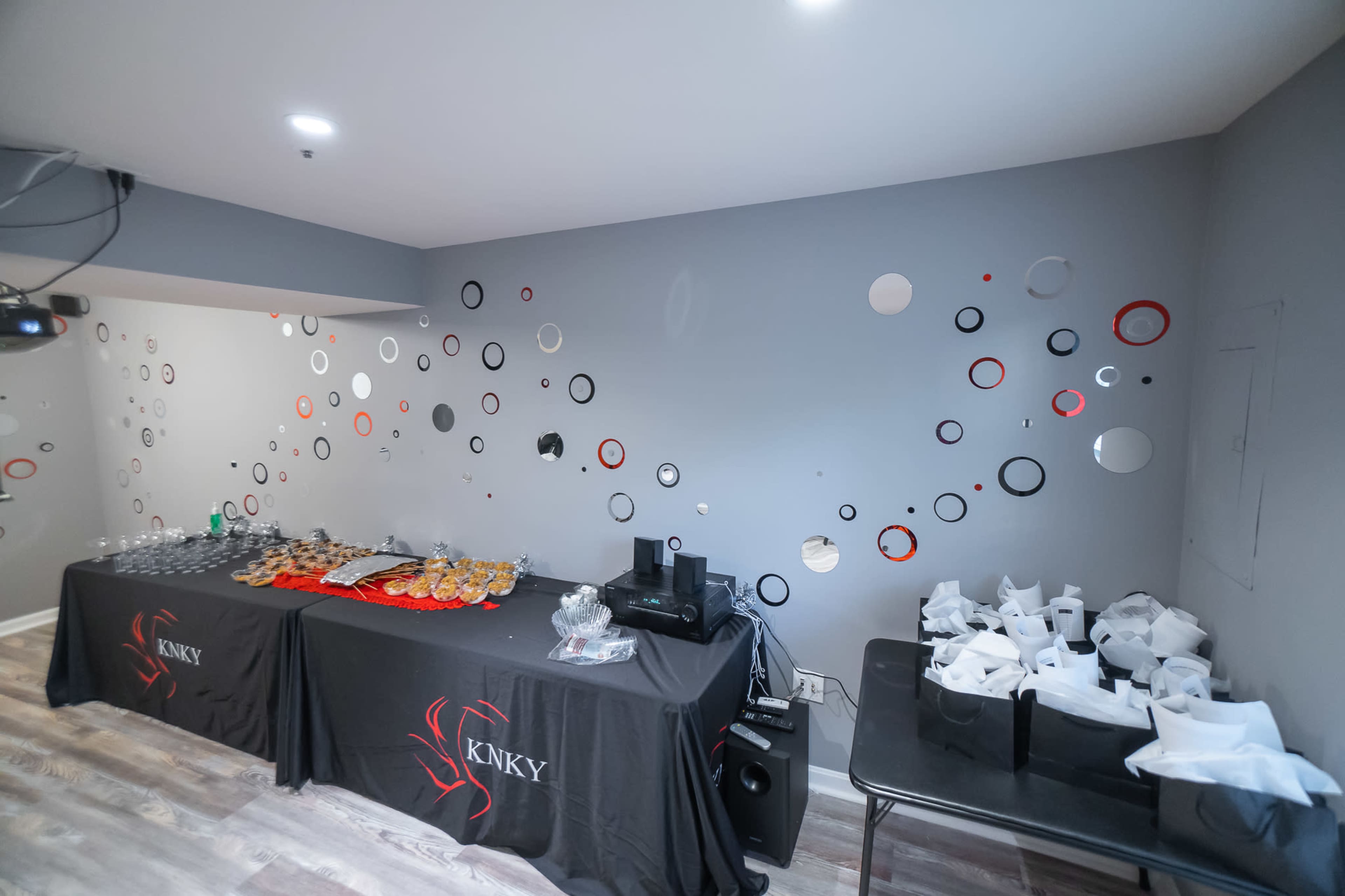 The image shows a room with two tables, one displaying various food items and the other with neatly arranged takeout bags, set against a wall decorated with circular wall art.