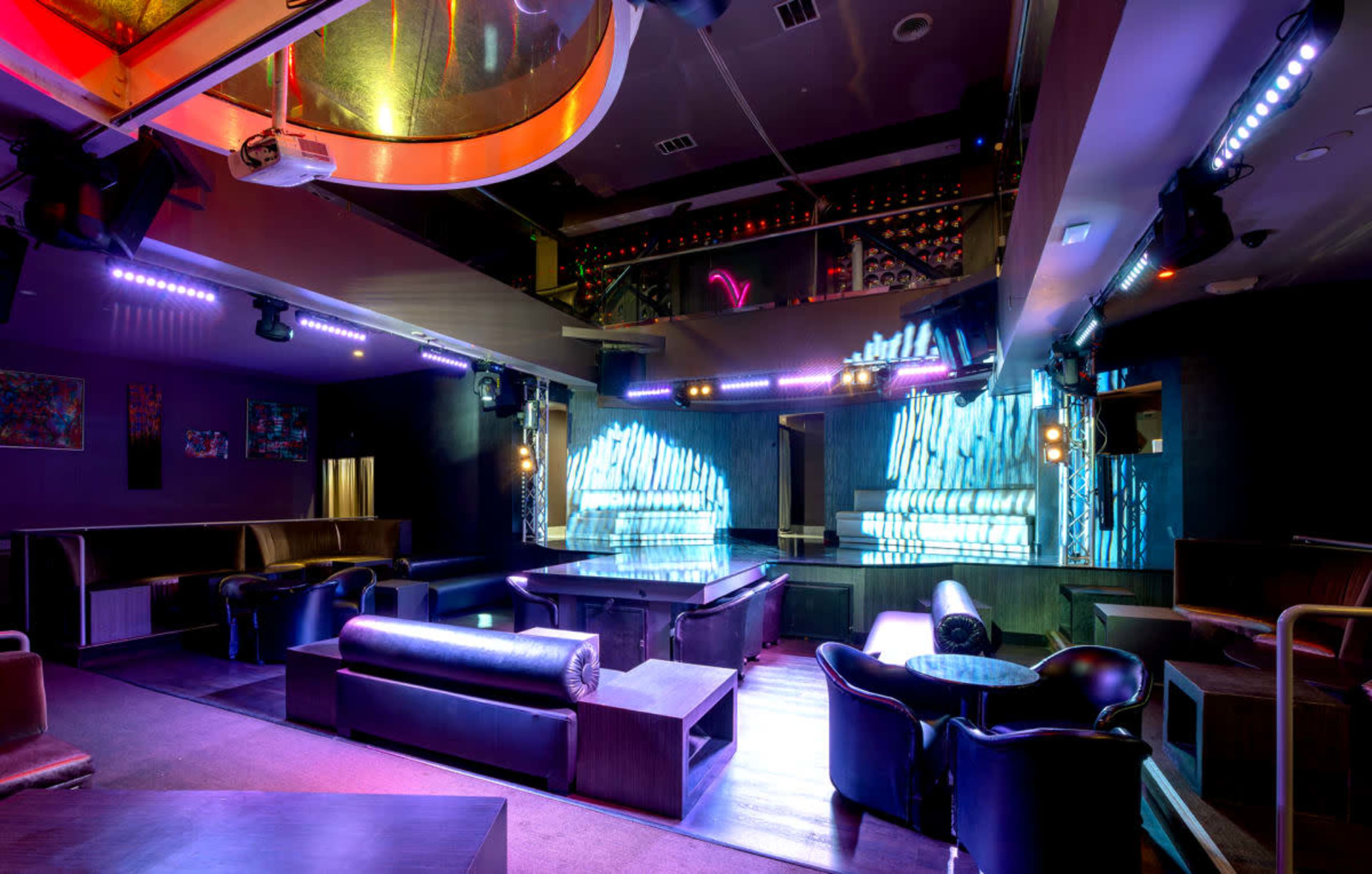 The image shows a modern nightclub interior with dark furnishings, a dance floor illuminated by colorful lights, and a bar area in the background.