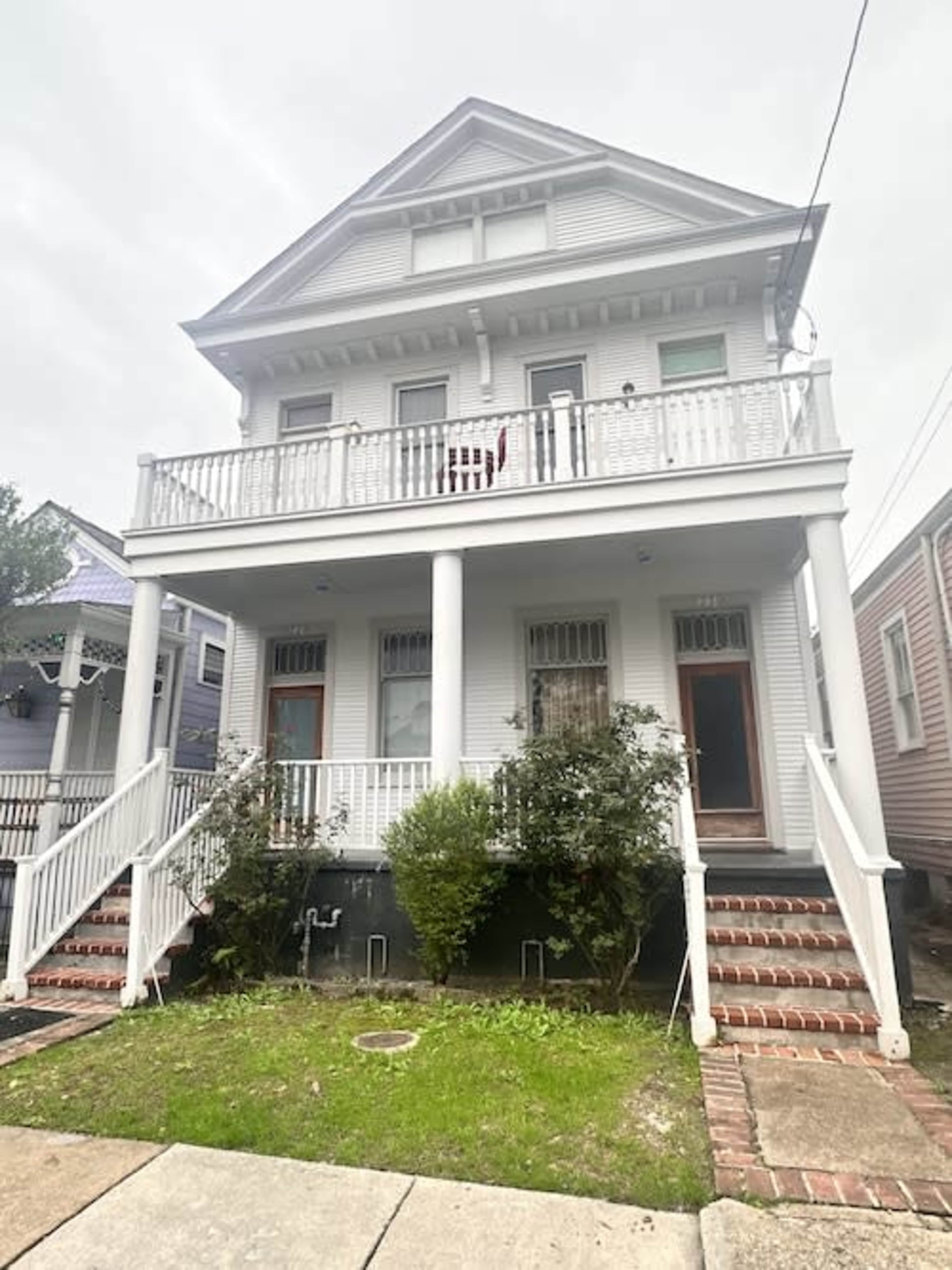 A two-story white house with a balcony and a front staircase stands among other similar homes.
