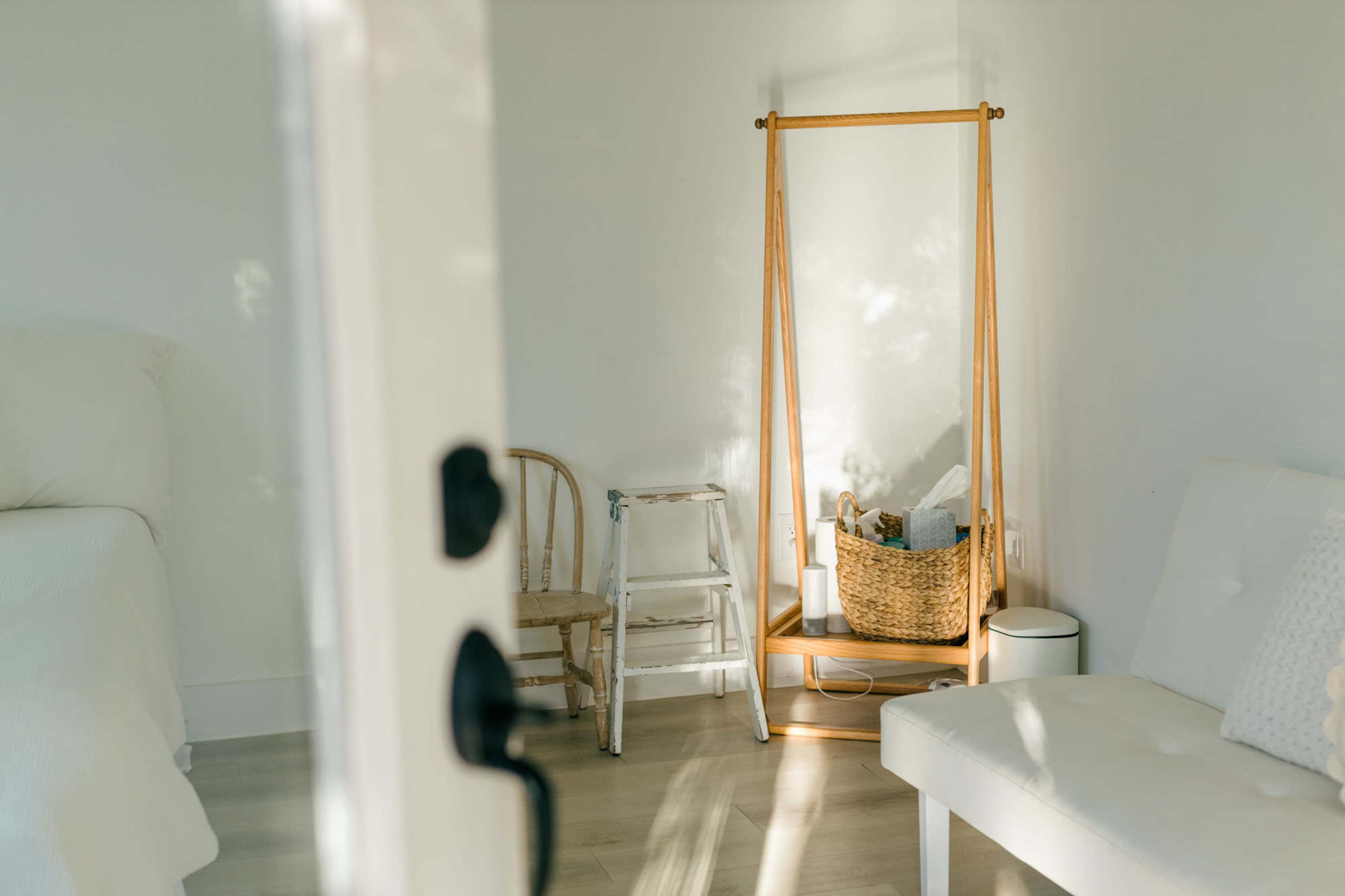 The image shows a bright, minimalist bedroom featuring a bed, a white couch, a wooden chair, and a tall wooden stand with a basket.