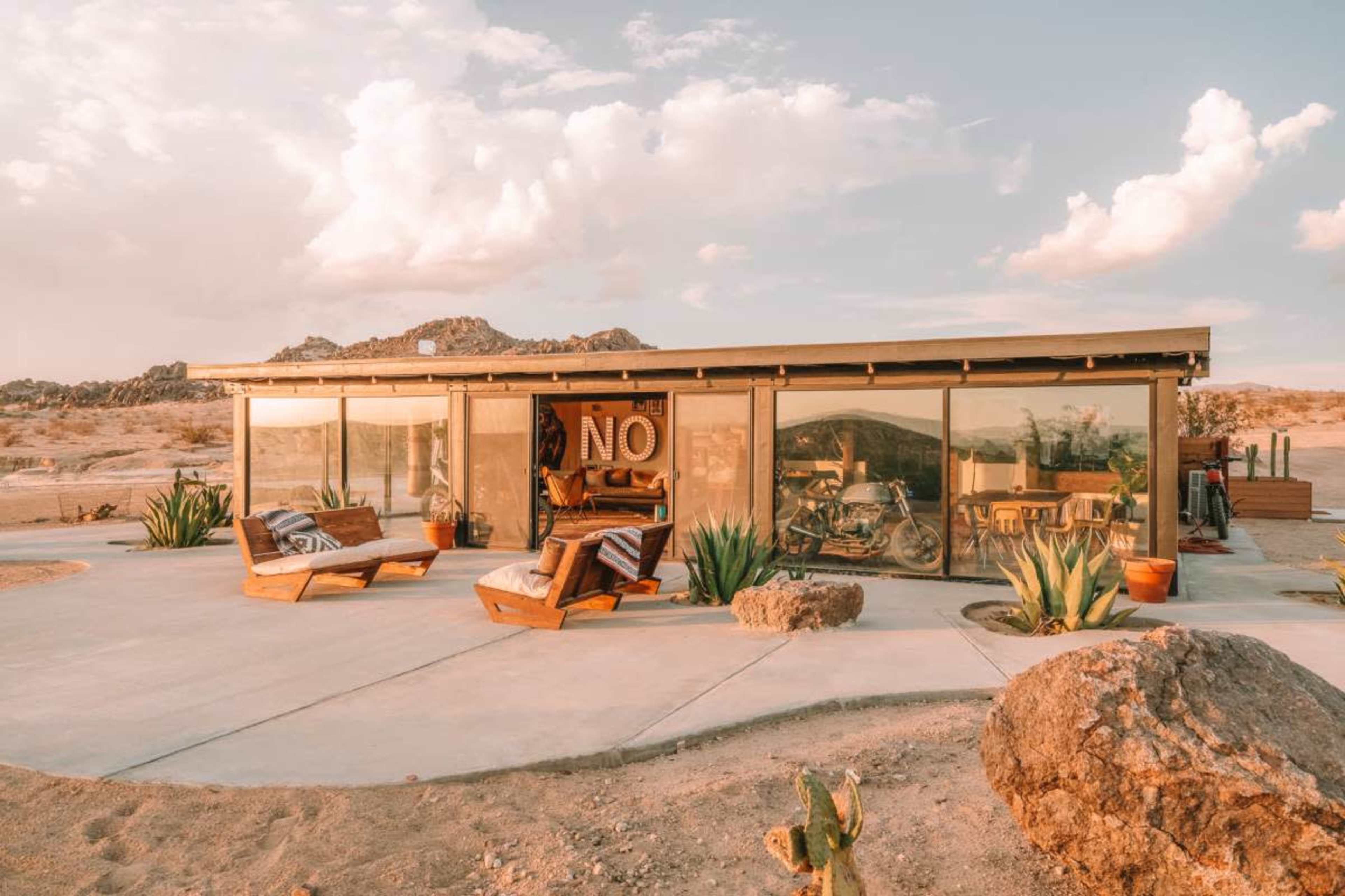 A modern glass-fronted house with wooden seating areas is situated in a desert landscape, surrounded by cacti and rocky formations.
