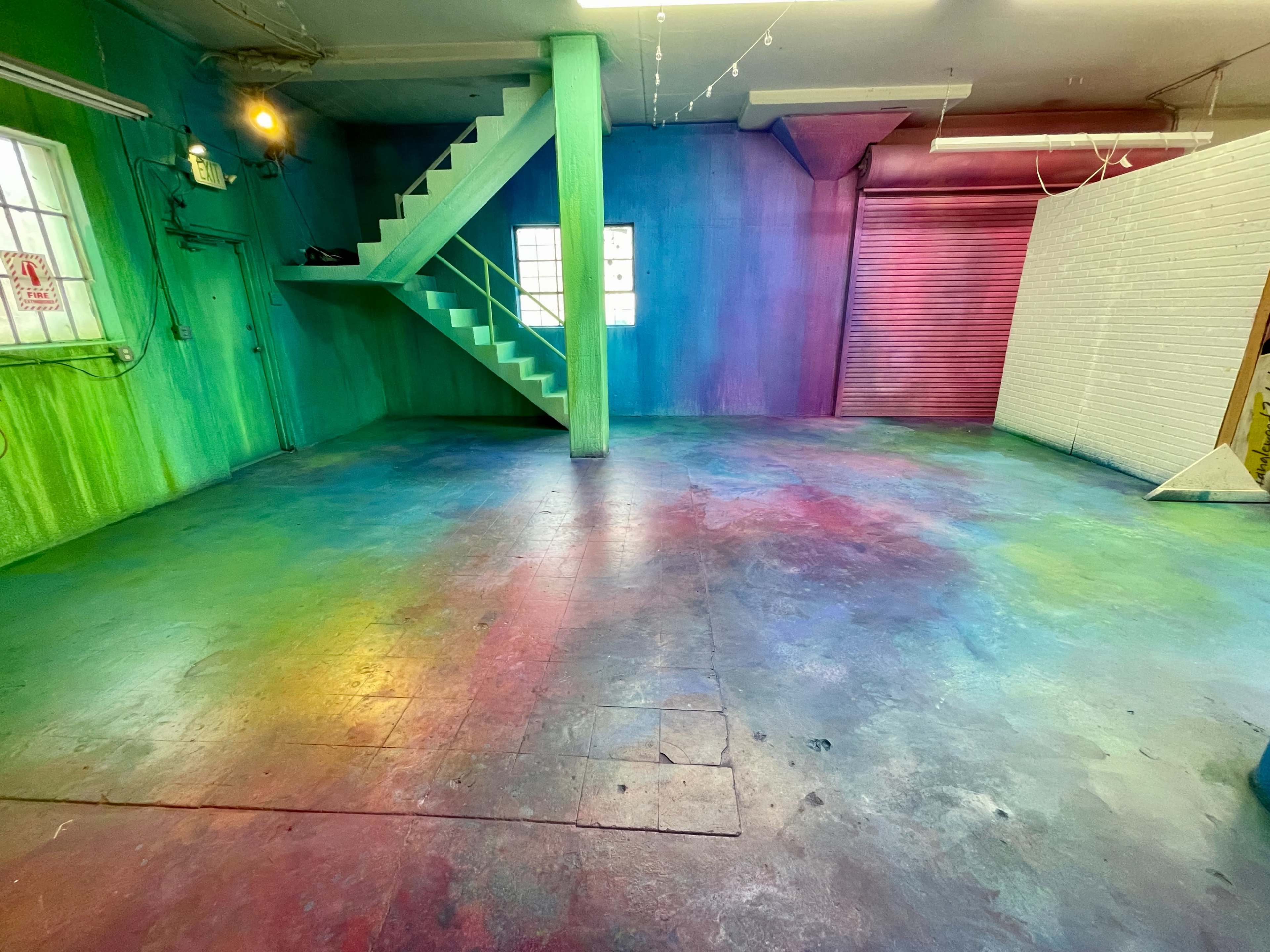 The image shows an interior space with colorful walls and floor, featuring a staircase on the left side and a partially closed shutter on the right.