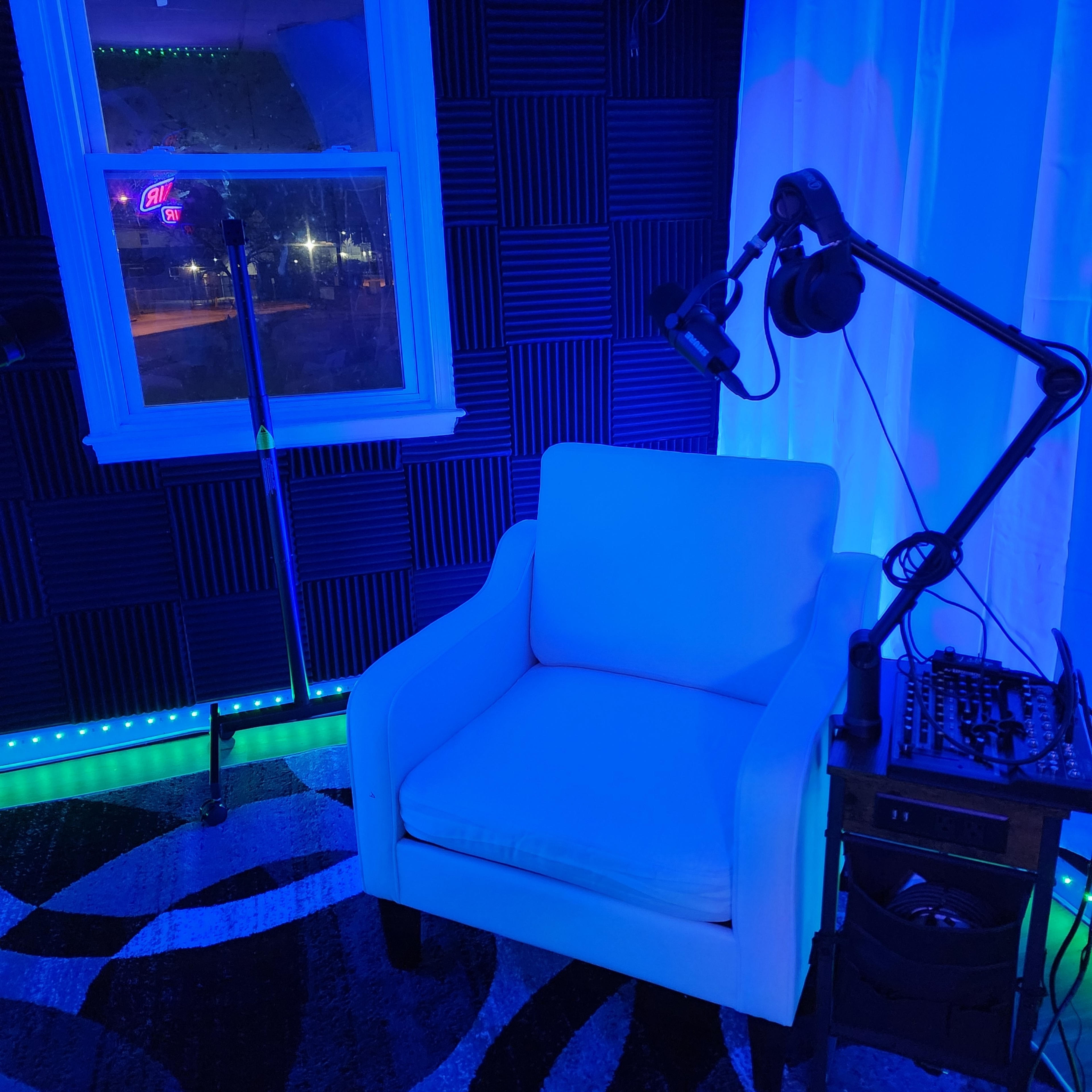 A recording studio corner with a white chair, a microphone on a stand, and headphones mounted on an adjustable arm, illuminated by blue lighting.