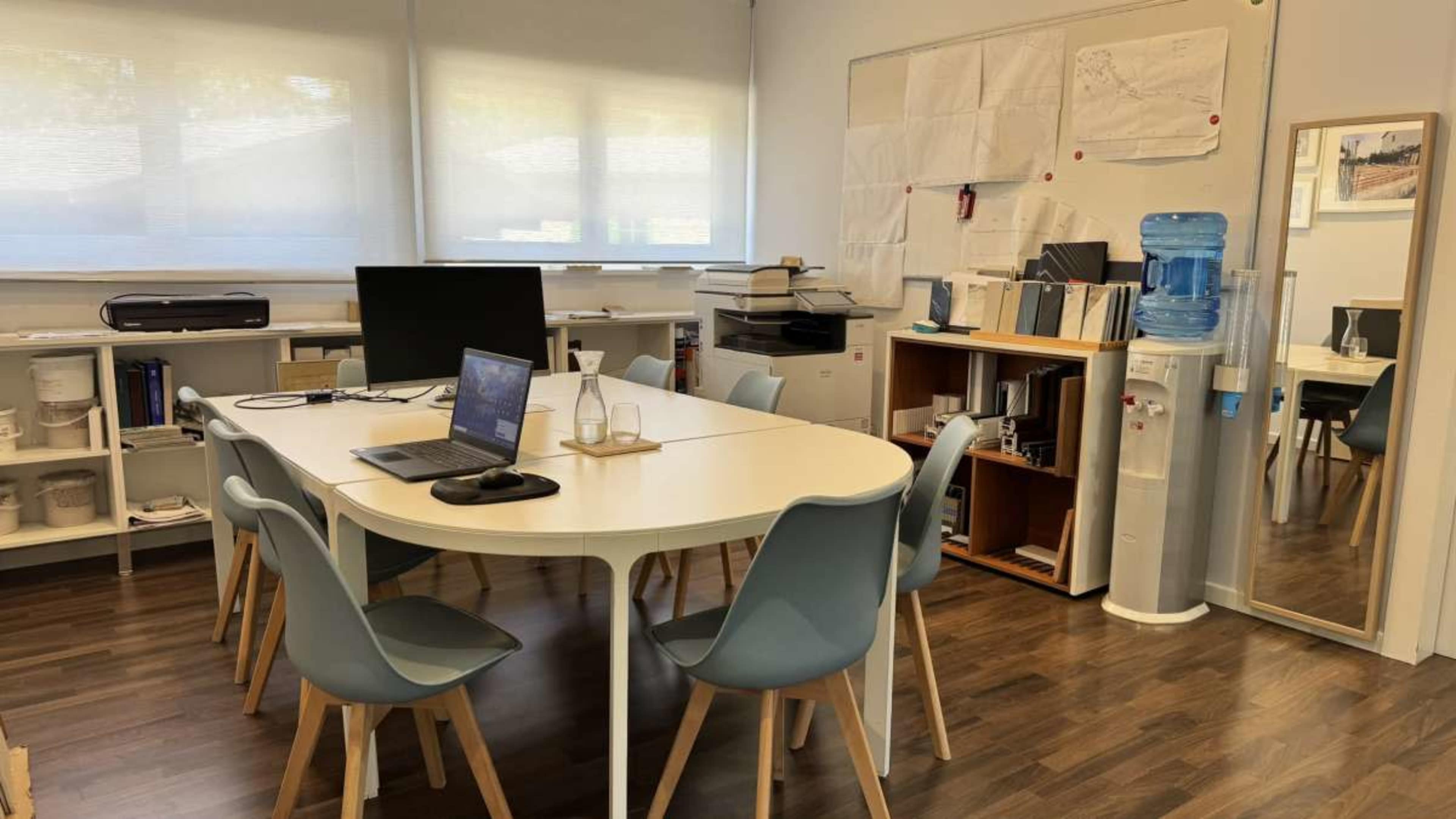 A modern, well-lit office meeting room features a large table surrounded by chairs, with a computer setup, a water dispenser, and various storage units against the walls.