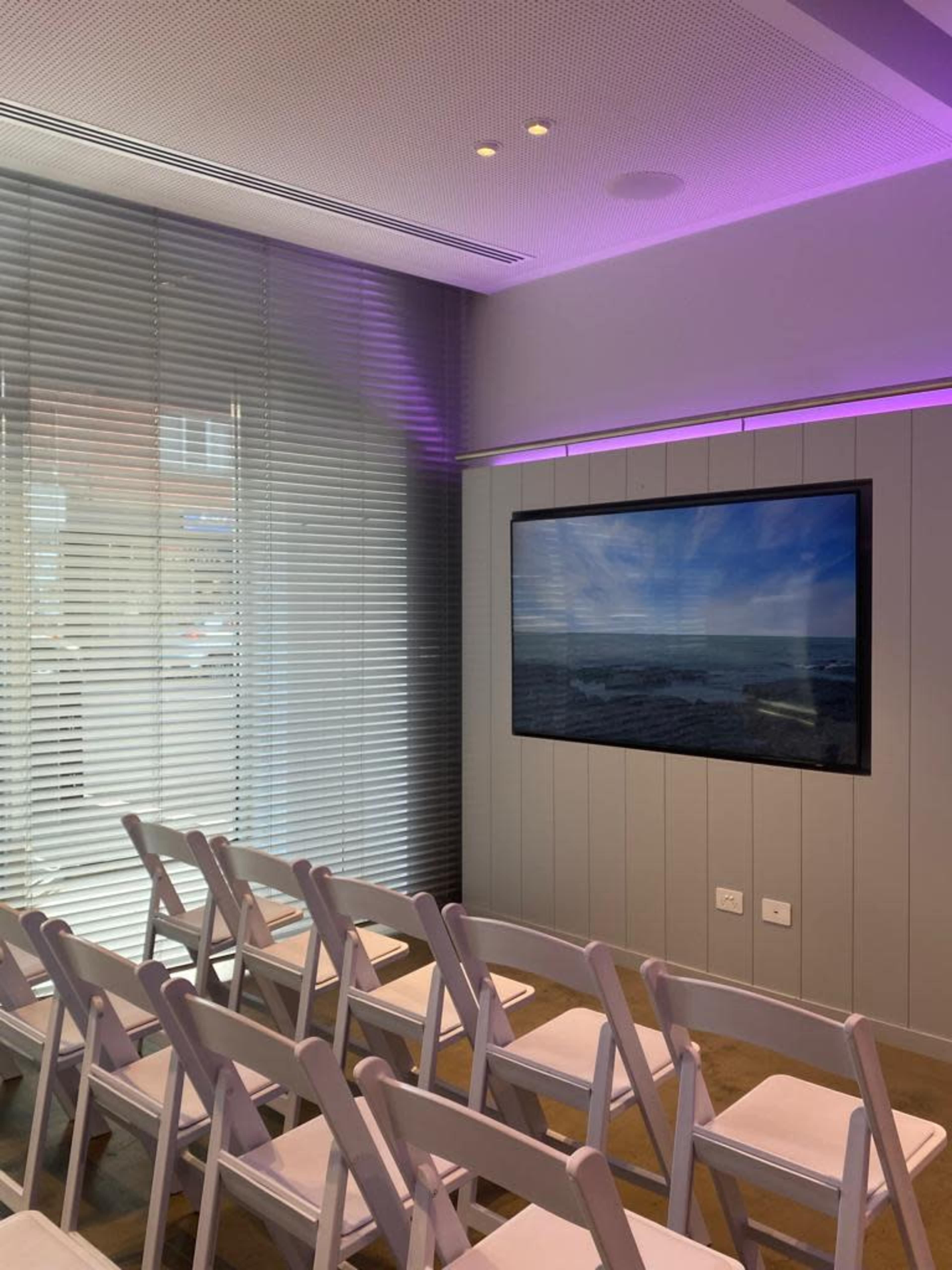 A modern meeting room features rows of white chairs facing a large screen mounted on the wall, with purple ambient lighting and window shades.