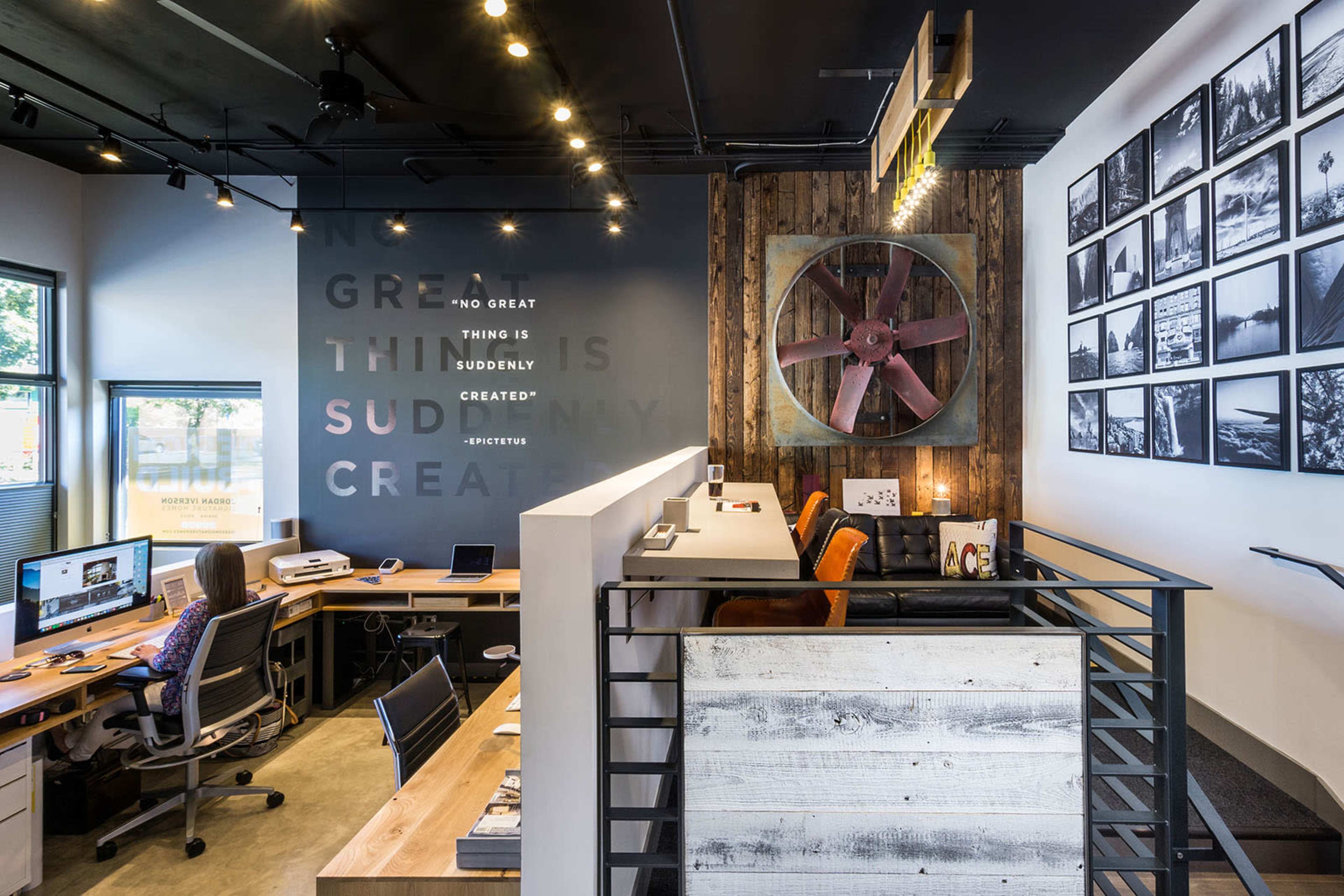 A modern office space with wooden desks, a large wall fan, a photo gallery, and a motivational quote on the wall.