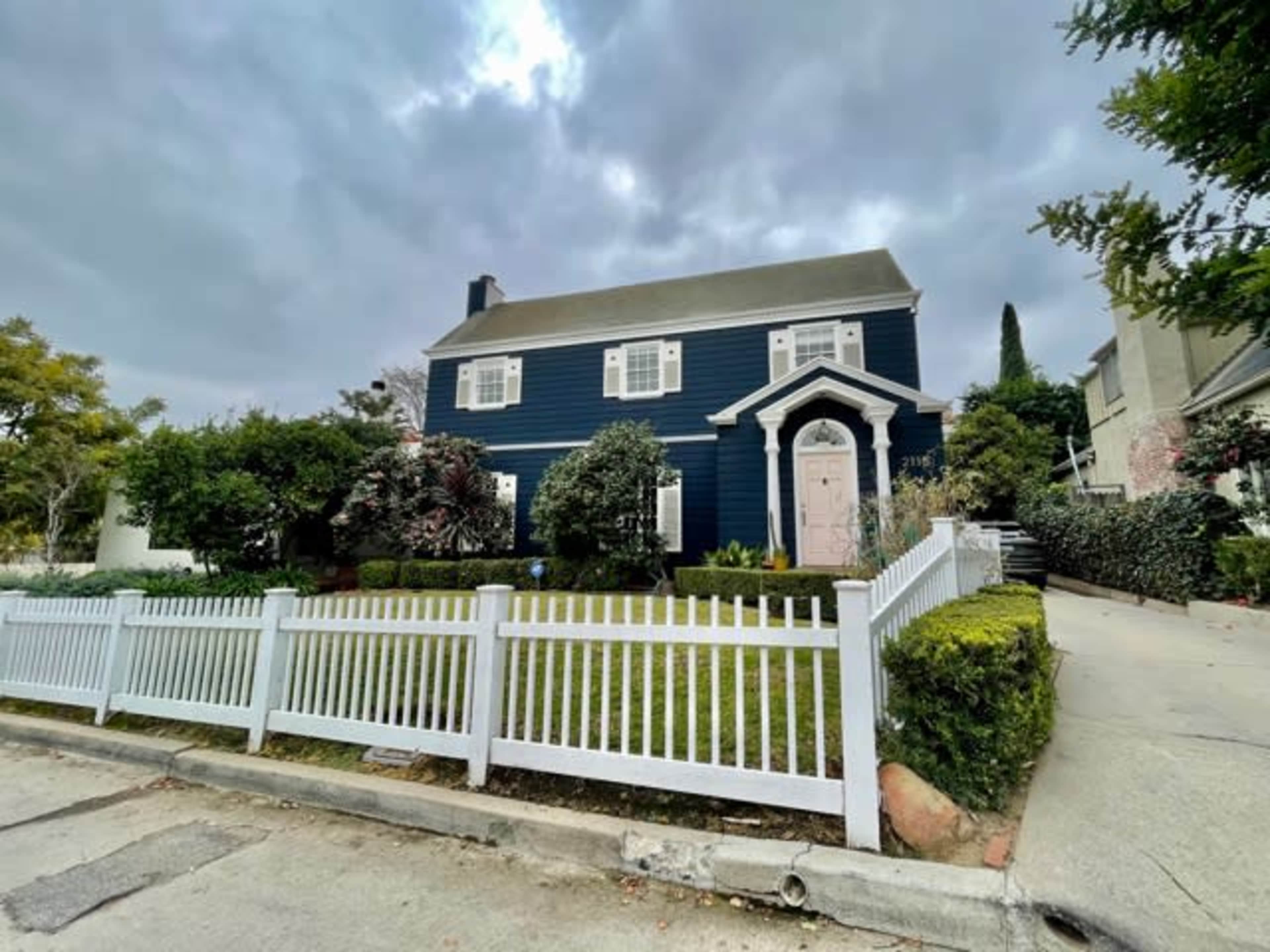 A two-story blue house with white trim and a pink door is surrounded by a white picket fence and a neatly maintained yard.