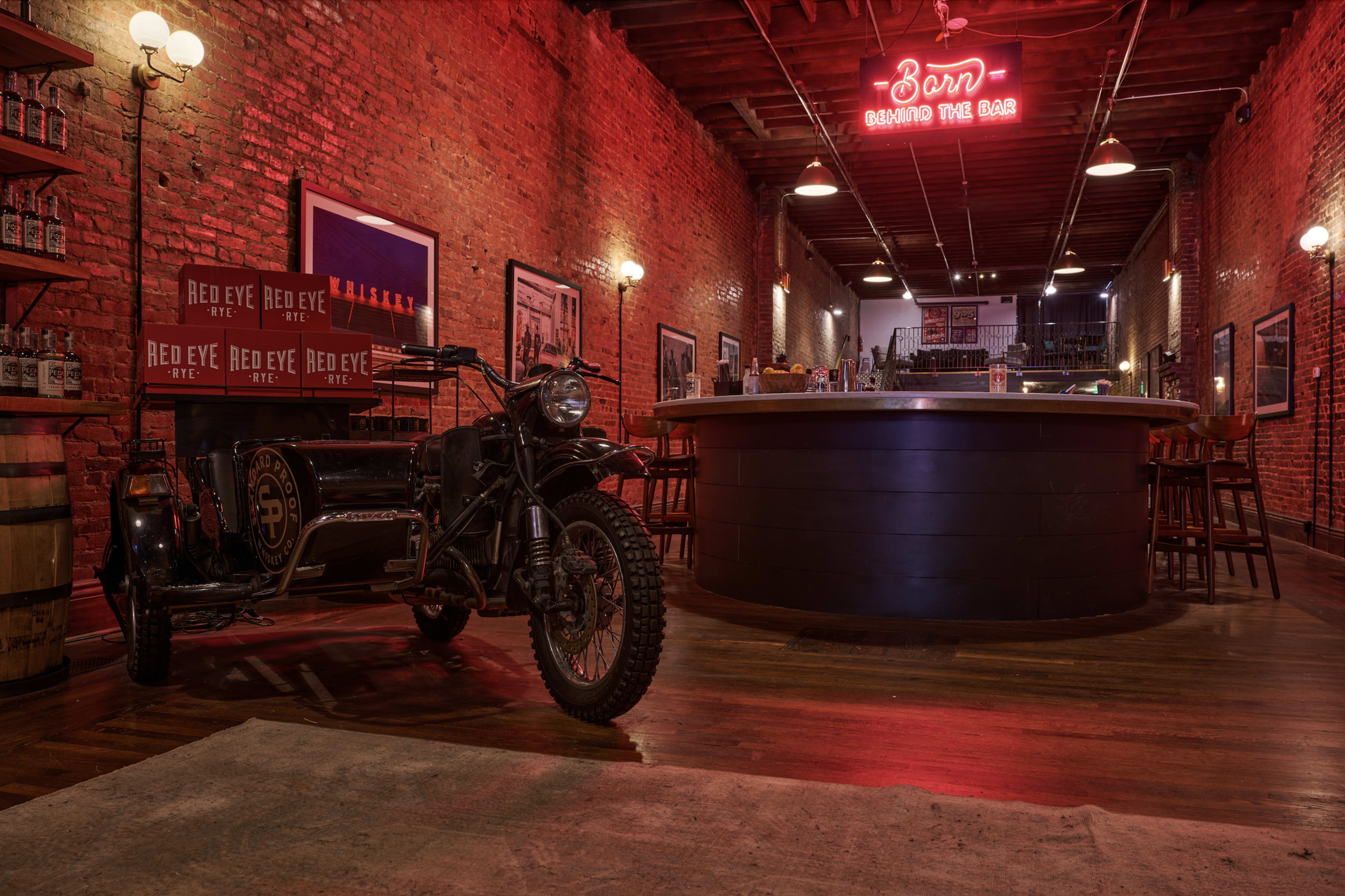 The image shows an interior of a bar with exposed brick walls, a circular bar counter, and a vintage motorcycle with a sidecar parked beside it.