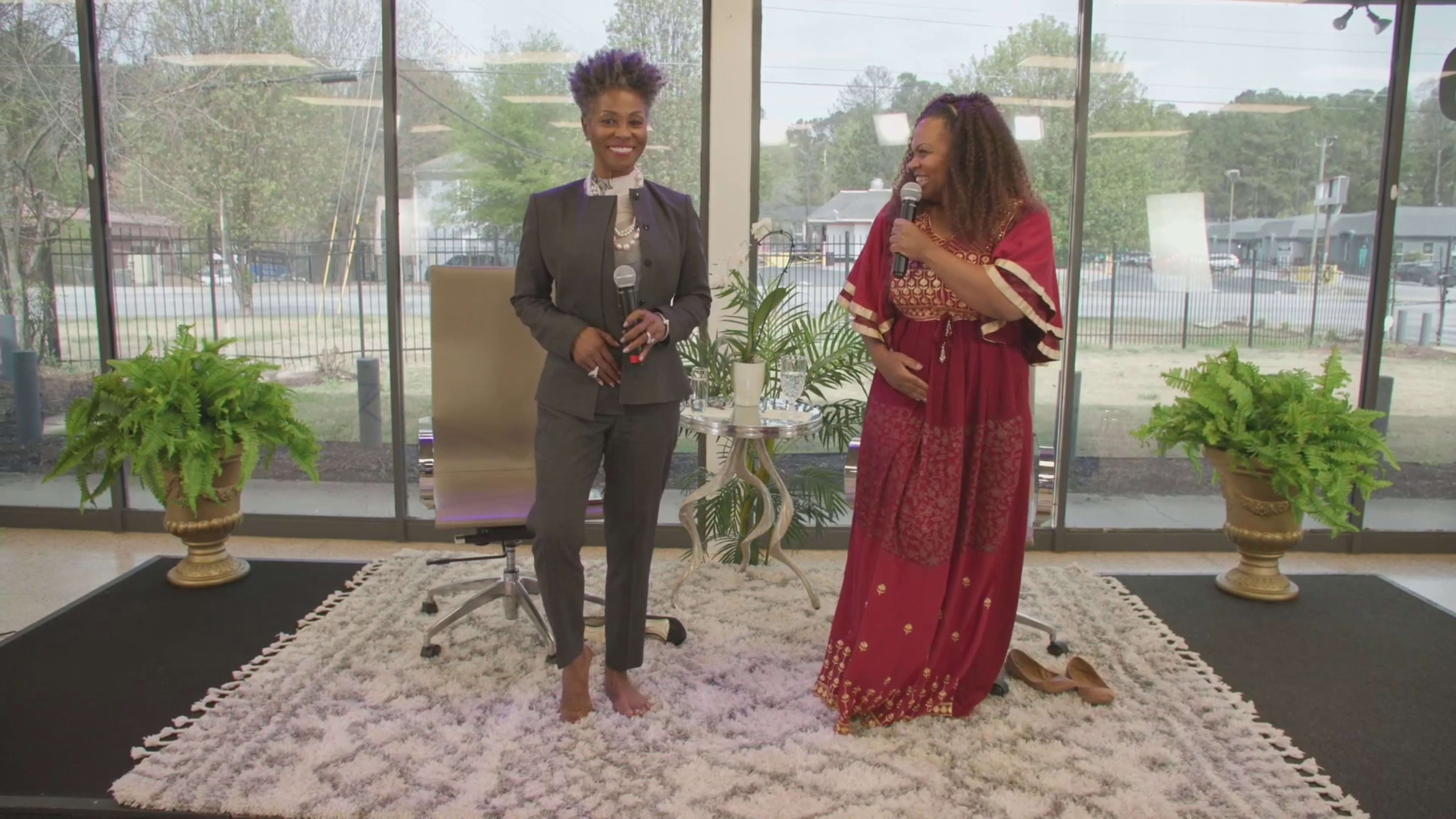 Two women stand together in a bright room with large windows, one dressed in a suit and the other in a traditional robe, both smiling and holding microphones.