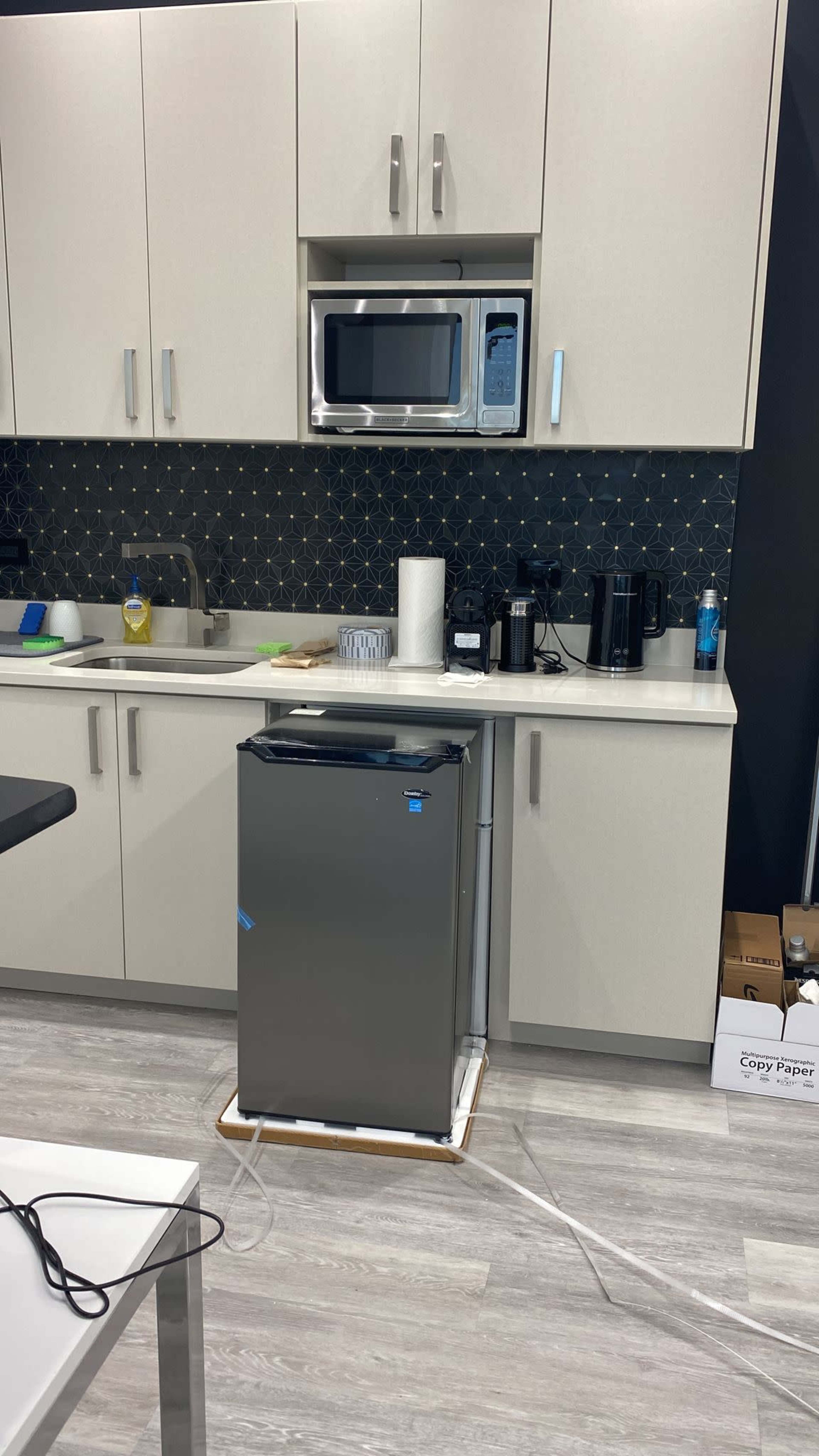A compact silver refrigerator stands on a wooden base in a modern kitchen with light-colored cabinets and a microwave above it.