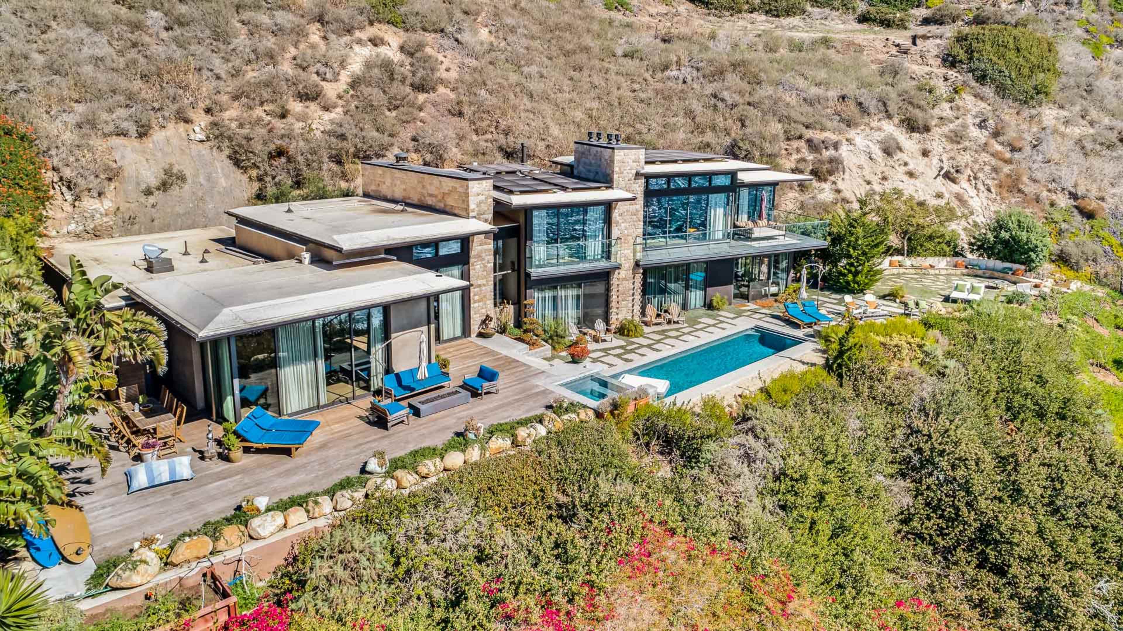 The image shows a modern hillside home with large glass windows, a swimming pool, and a landscaped garden.
