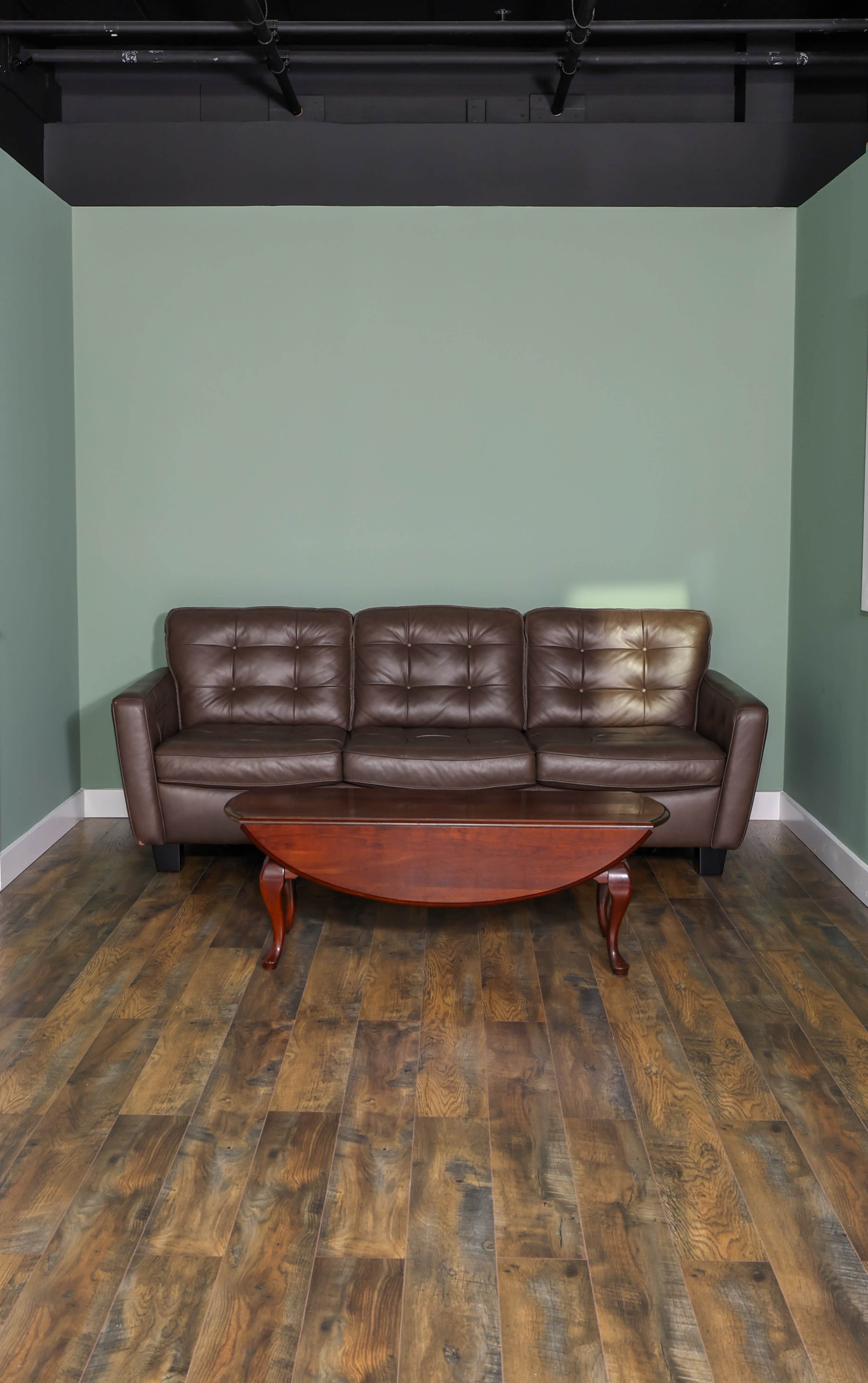 A brown leather sofa is positioned against a green wall, with a round wooden coffee table in front of it on a wooden floor.