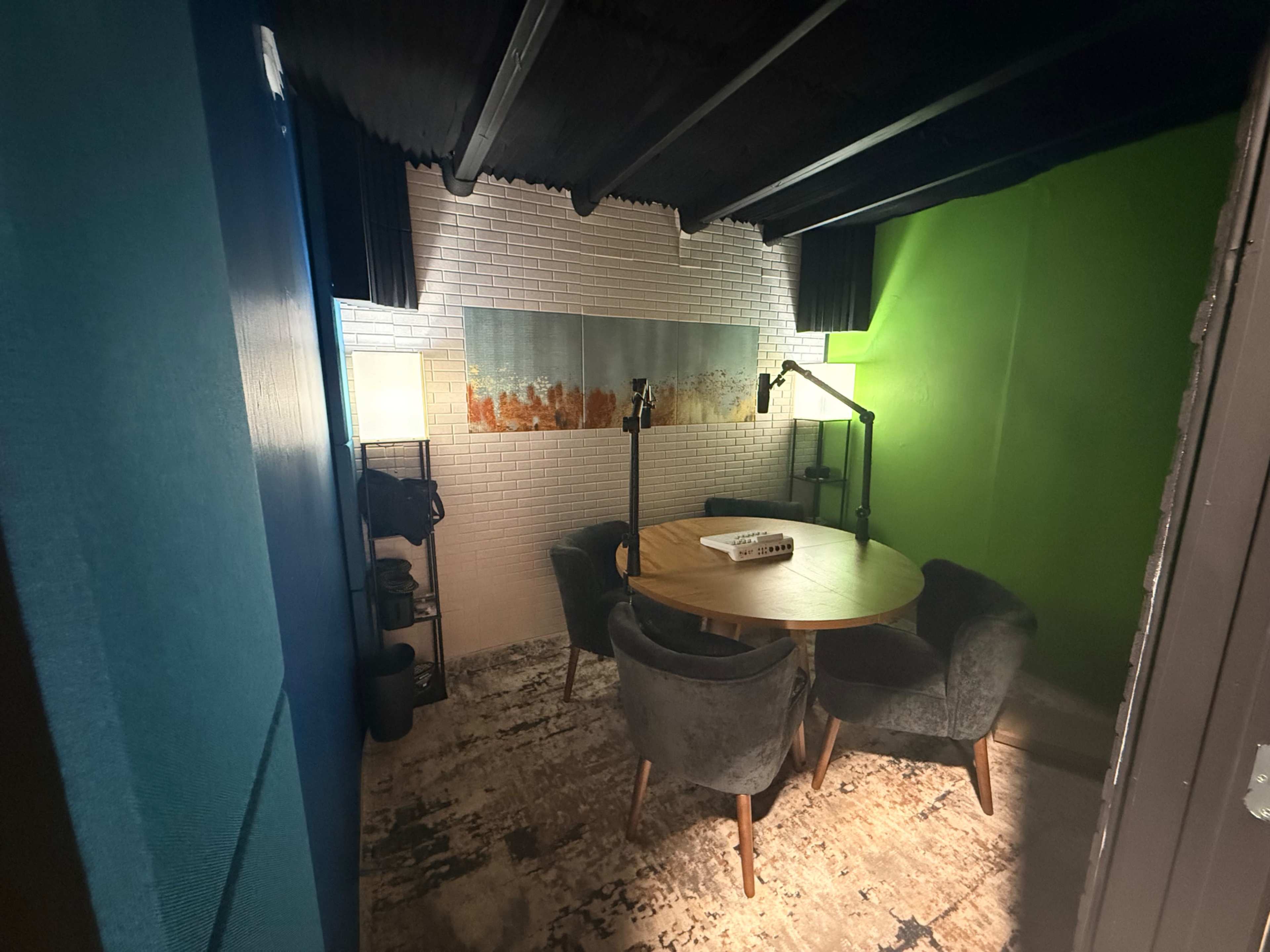 The image shows a small meeting room featuring a round wooden table surrounded by four upholstered chairs, with a colorful wall and a microphone setup for recording or presentations.