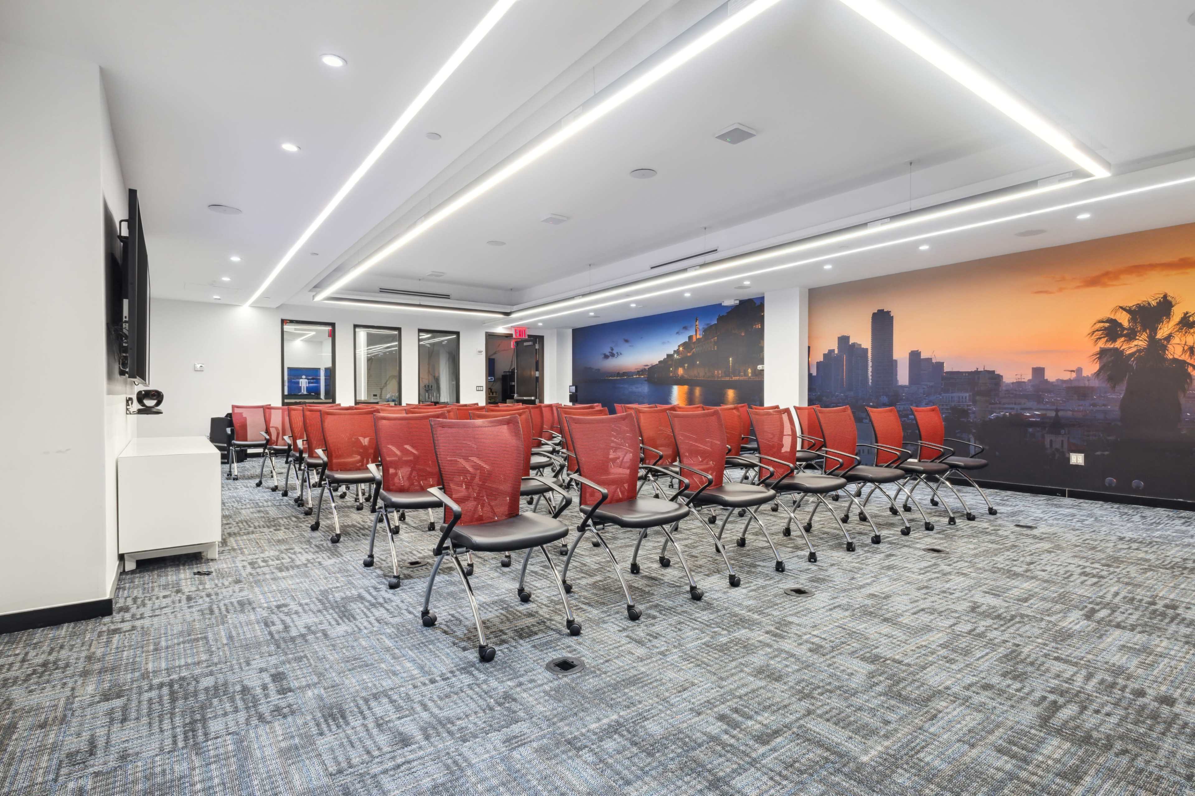 A modern conference room features rows of red chairs arranged facing a wall with a sunset cityscape mural.