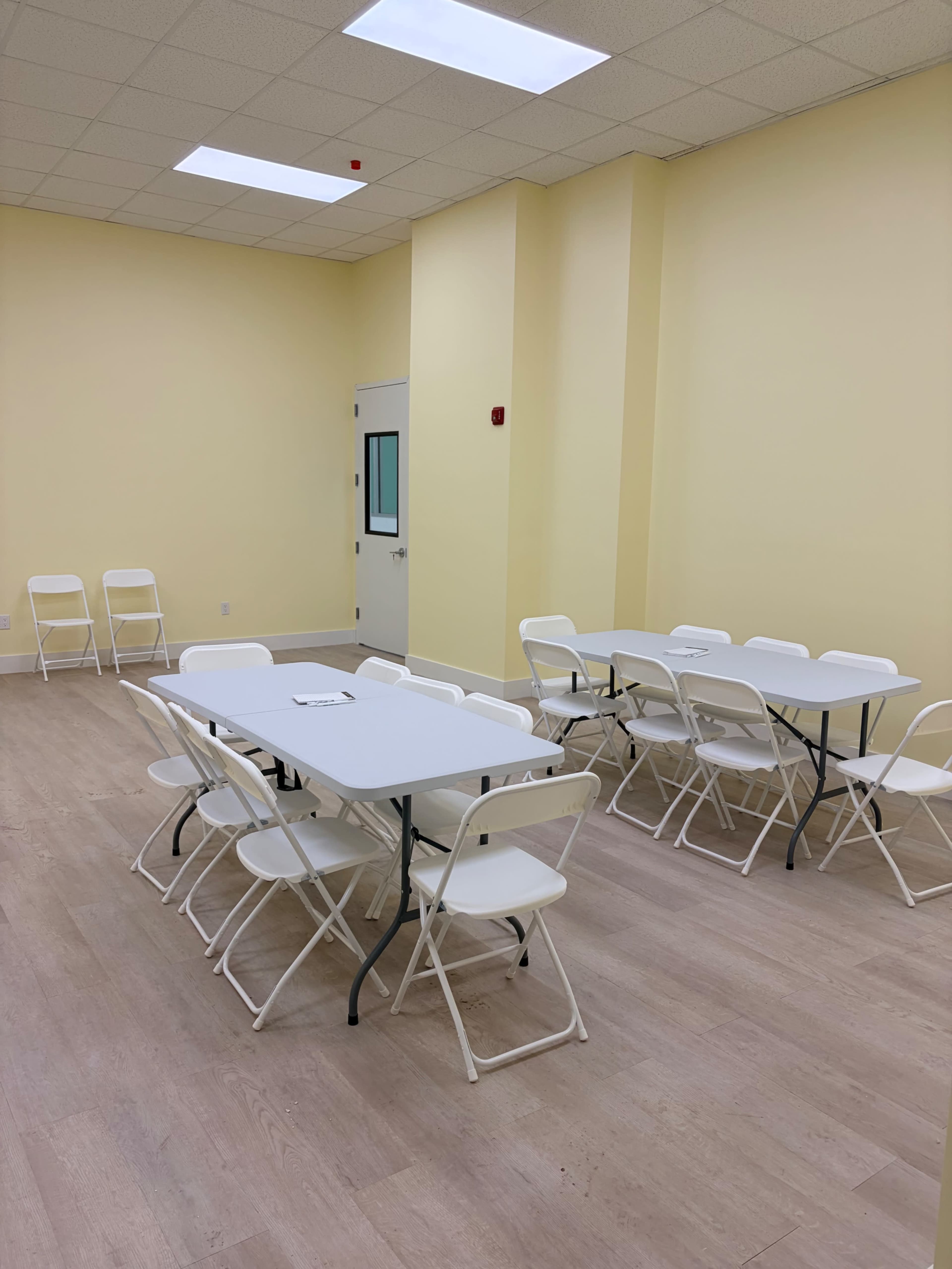 The image shows a bright, empty room with pale yellow walls filled with several white folding tables and chairs arranged neatly.