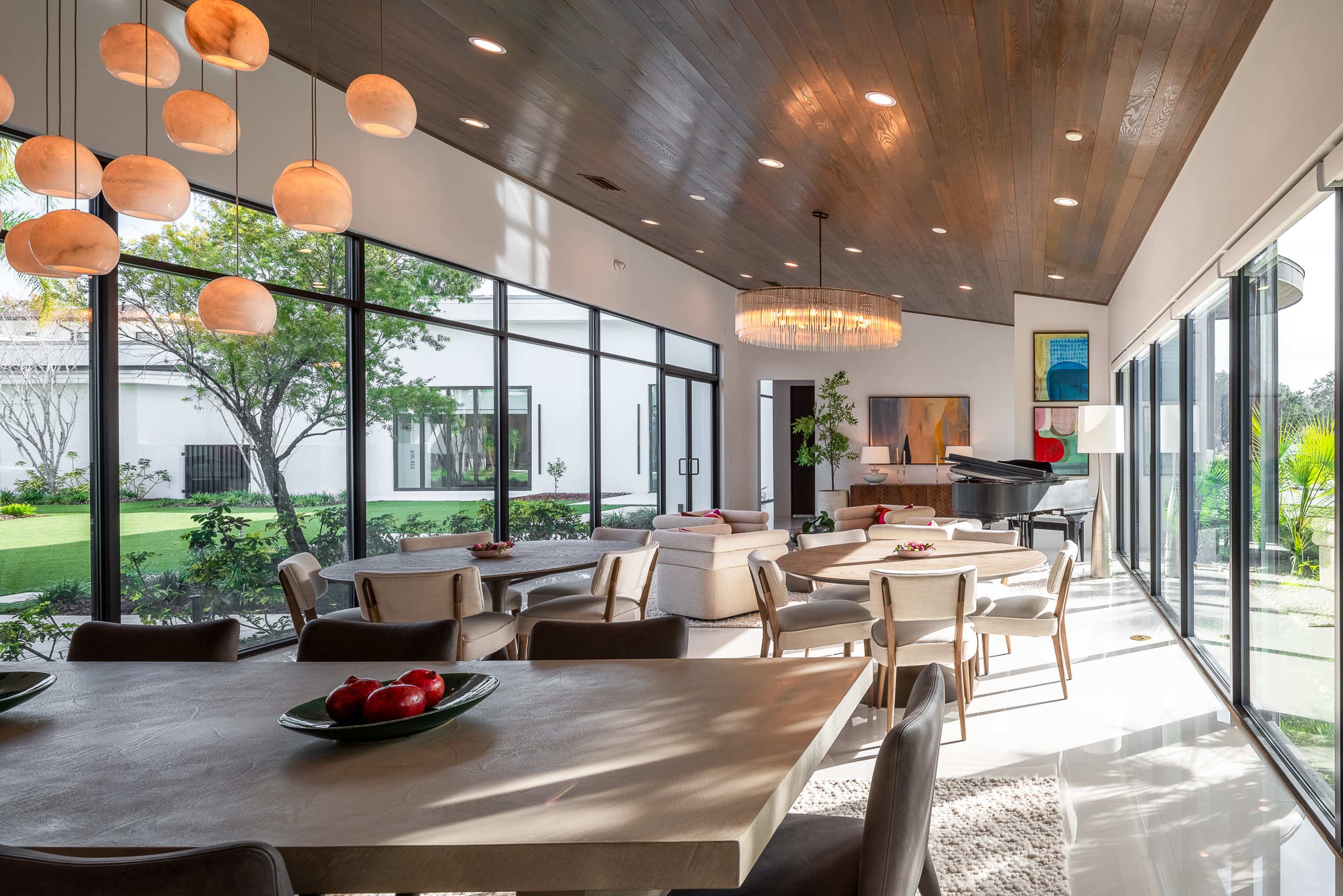 A modern dining area features a large table, surrounded by chairs, with floor-to-ceiling windows showcasing an outdoor garden and a grand piano in the corner.