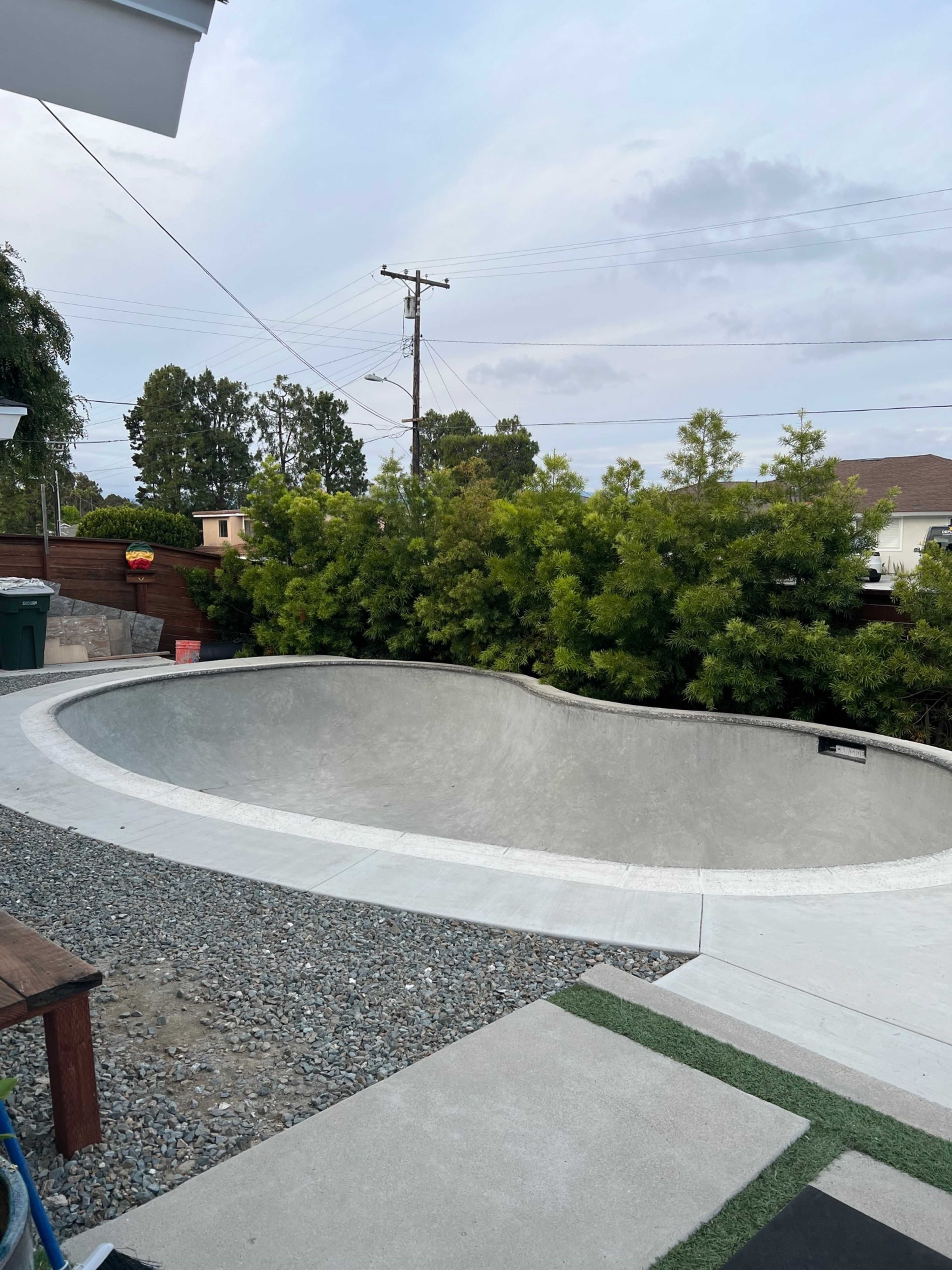 A concrete skate bowl located in a backyard, surrounded by greenery and a gravel area.