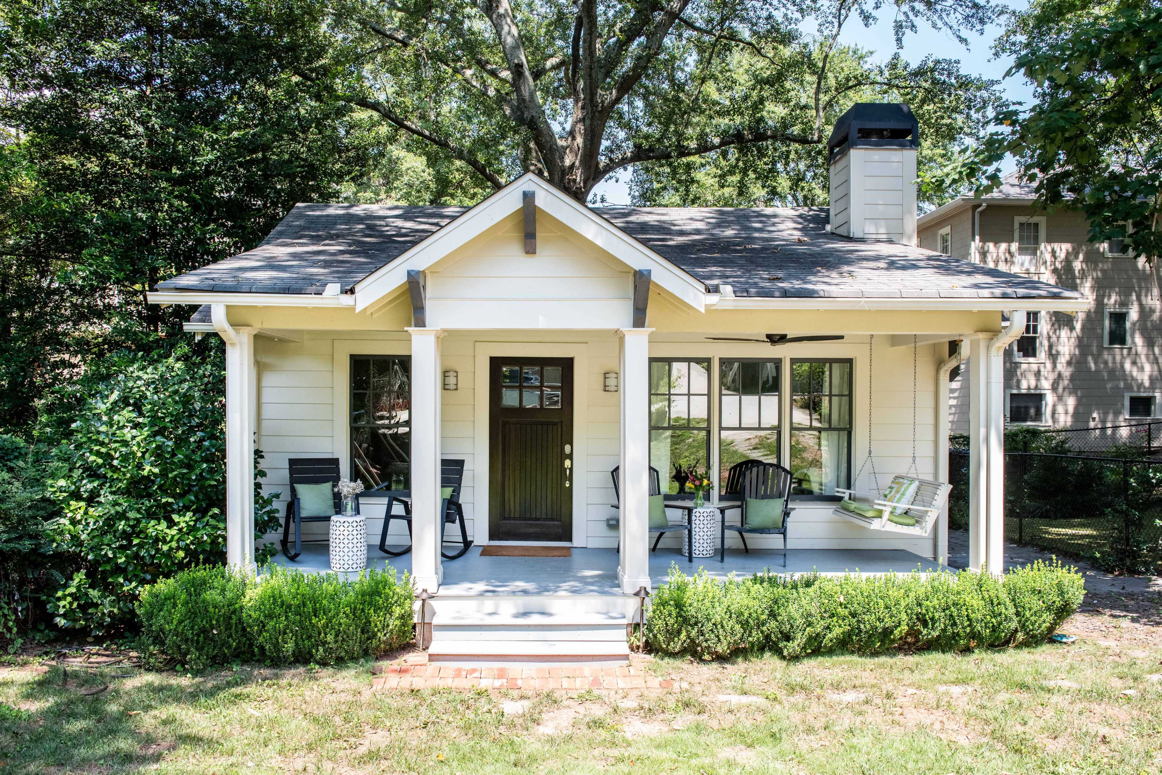 A small, white cottage with a front porch and green shrubs is surrounded by trees and a grassy lawn.