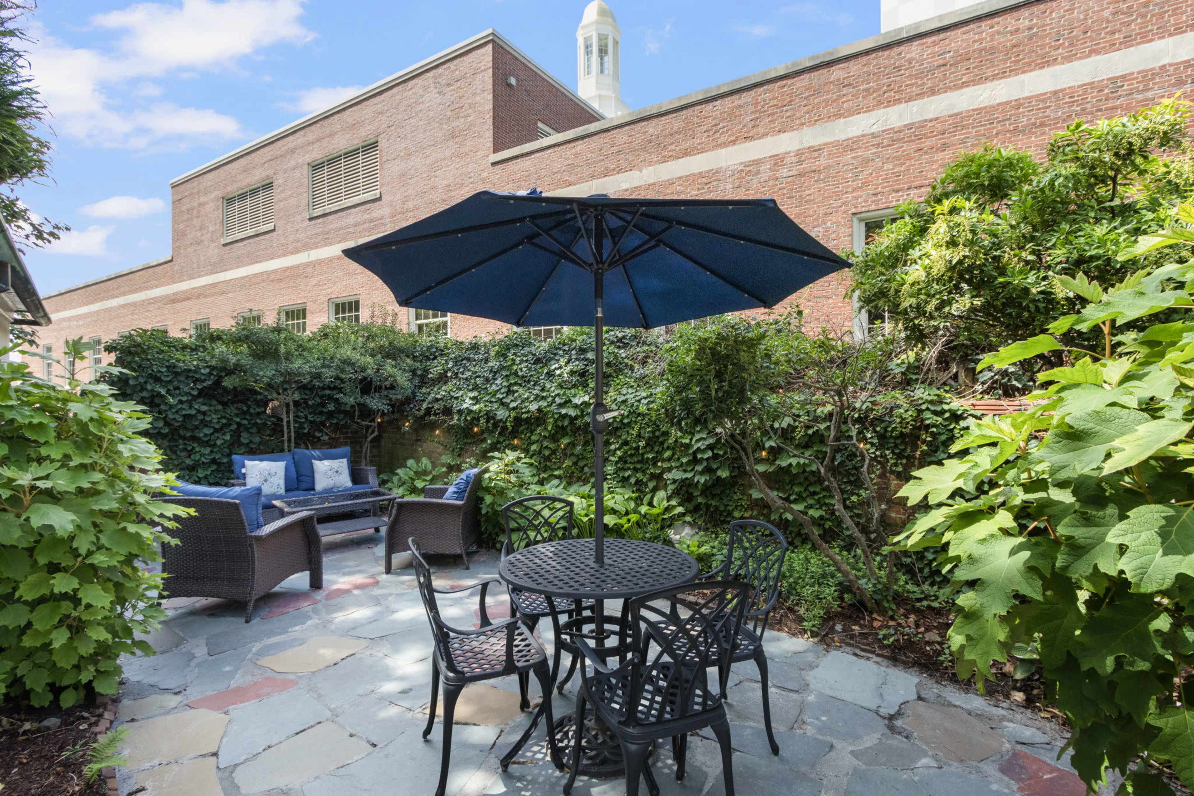 The image shows a small, landscaped outdoor patio area featuring a table with four chairs, an umbrella, and a seating arrangement surrounded by greenery and brick walls.