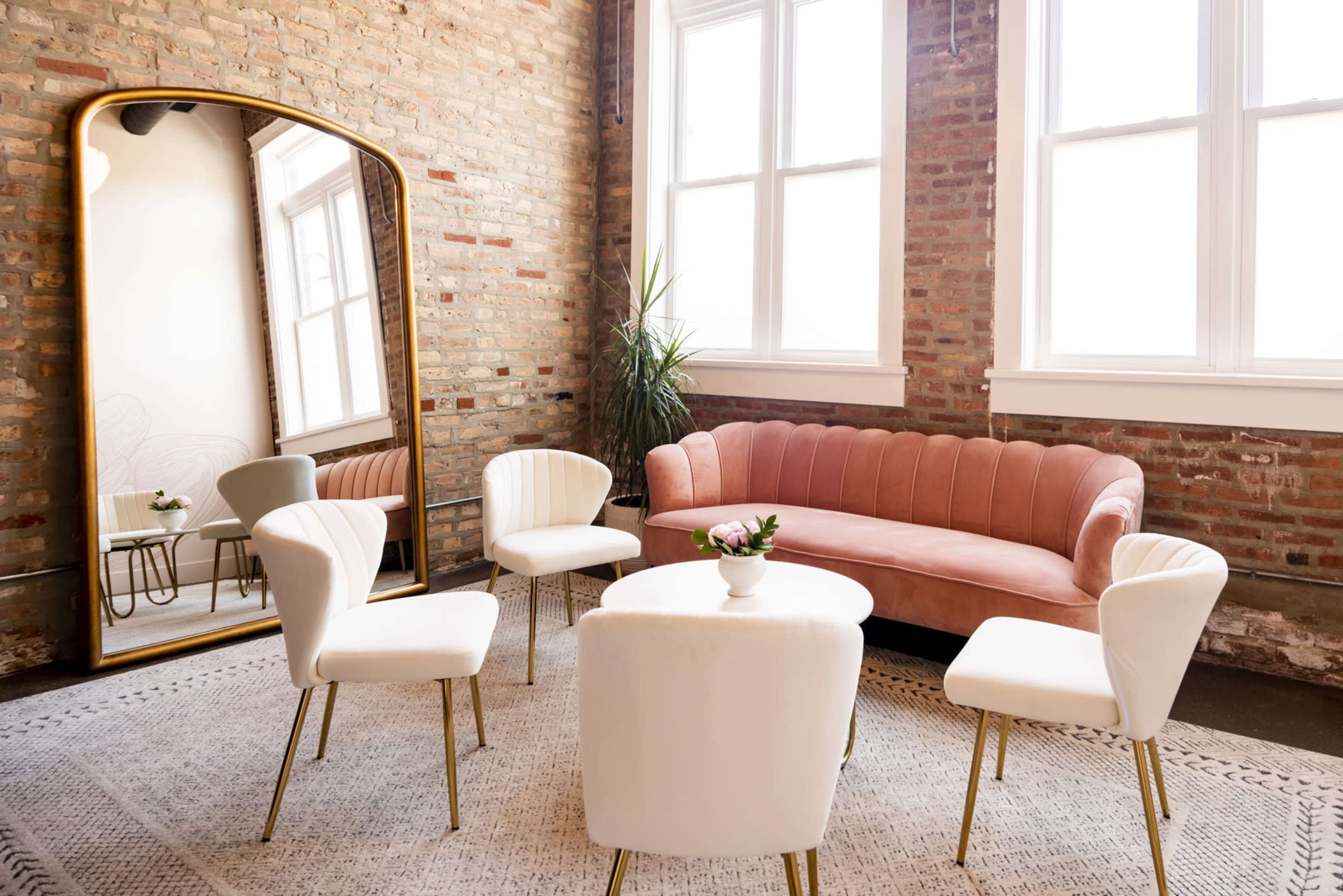A cozy seating area features a pink couch, four white chairs, and a round table, all set against a backdrop of exposed brick walls and large windows.
