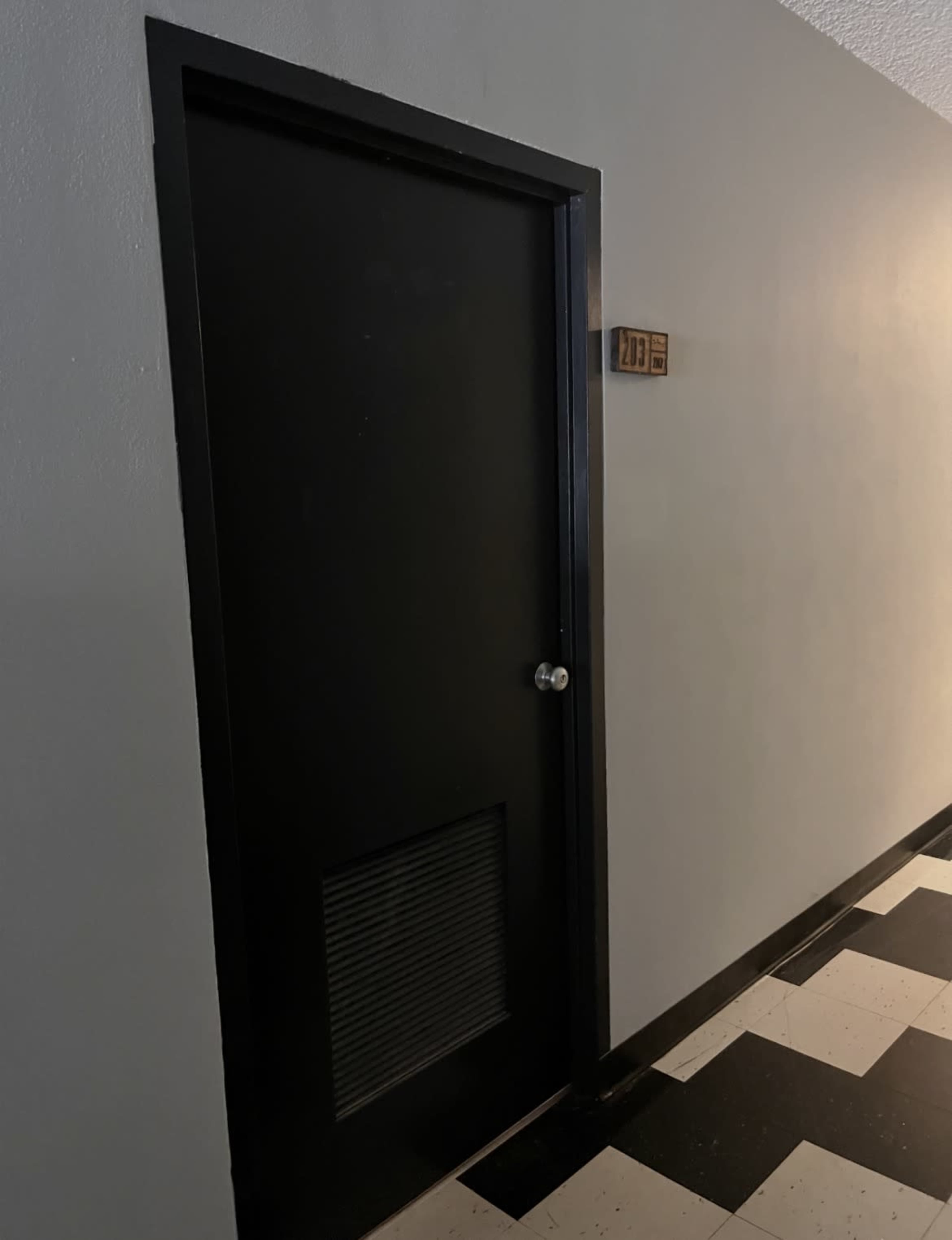 The image shows a black door with a vent at the lower part, located in a hallway with a pattern of black and white tiles.