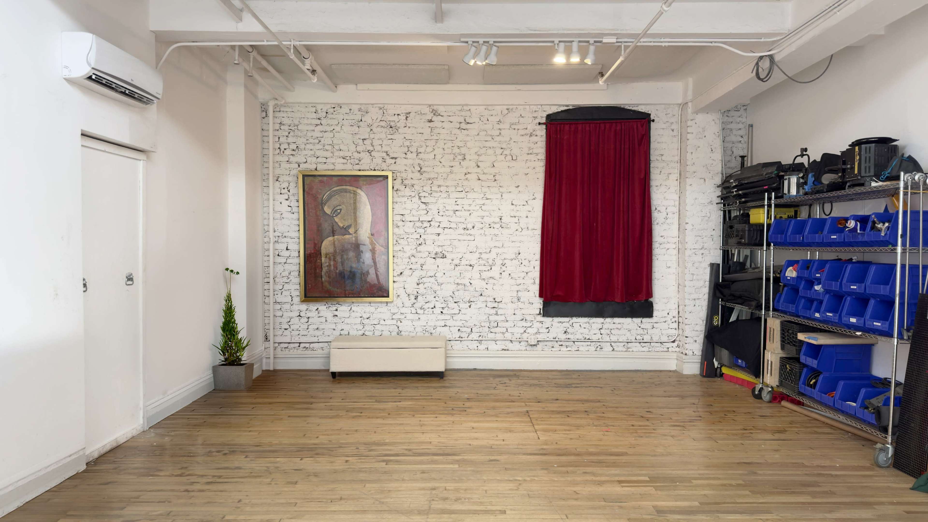 The image shows an empty room with a wooden floor, a brick wall adorned with a framed painting, a red curtain, and storage on the right side.