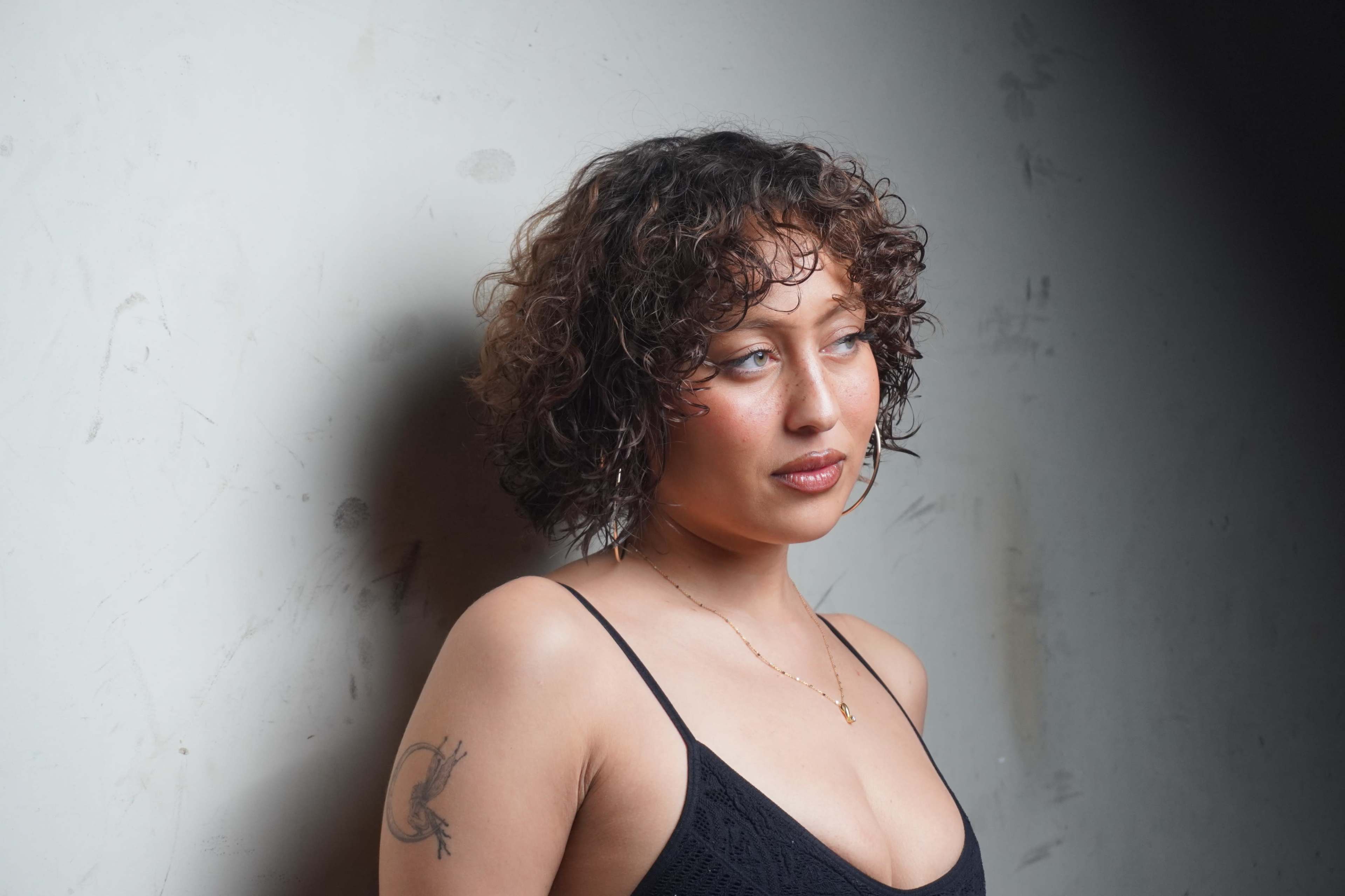 A woman with curly hair and a tattoo stands against a textured wall.