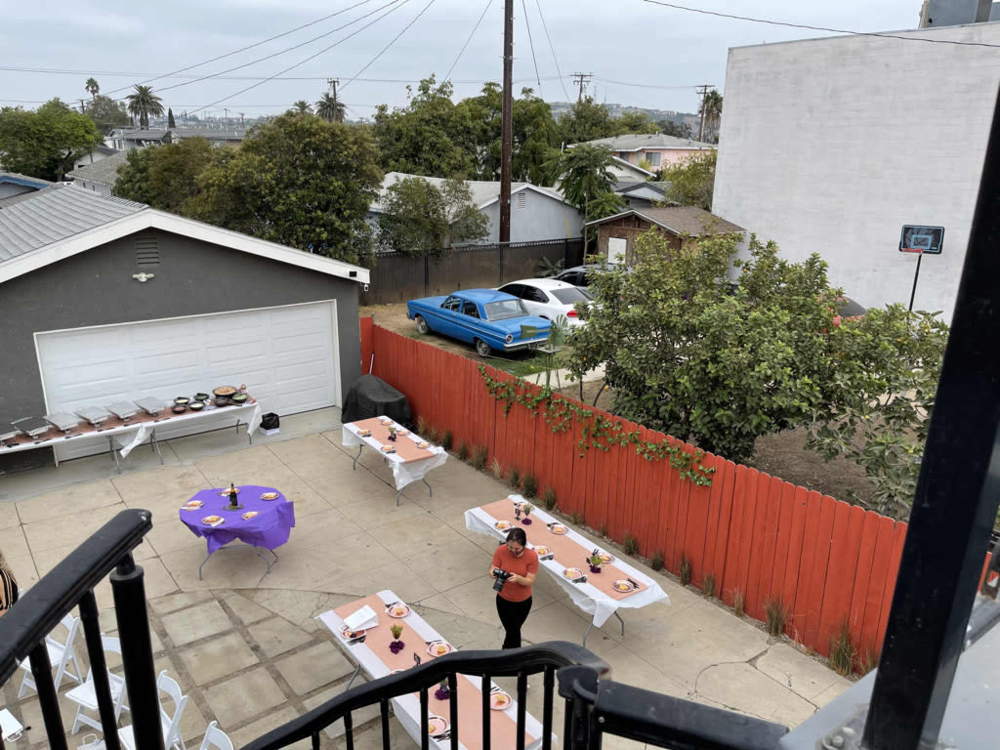The image shows a backyard setup for an event, featuring tables arranged with white tablecloths and purple decorations, and a blue car parked in the background.