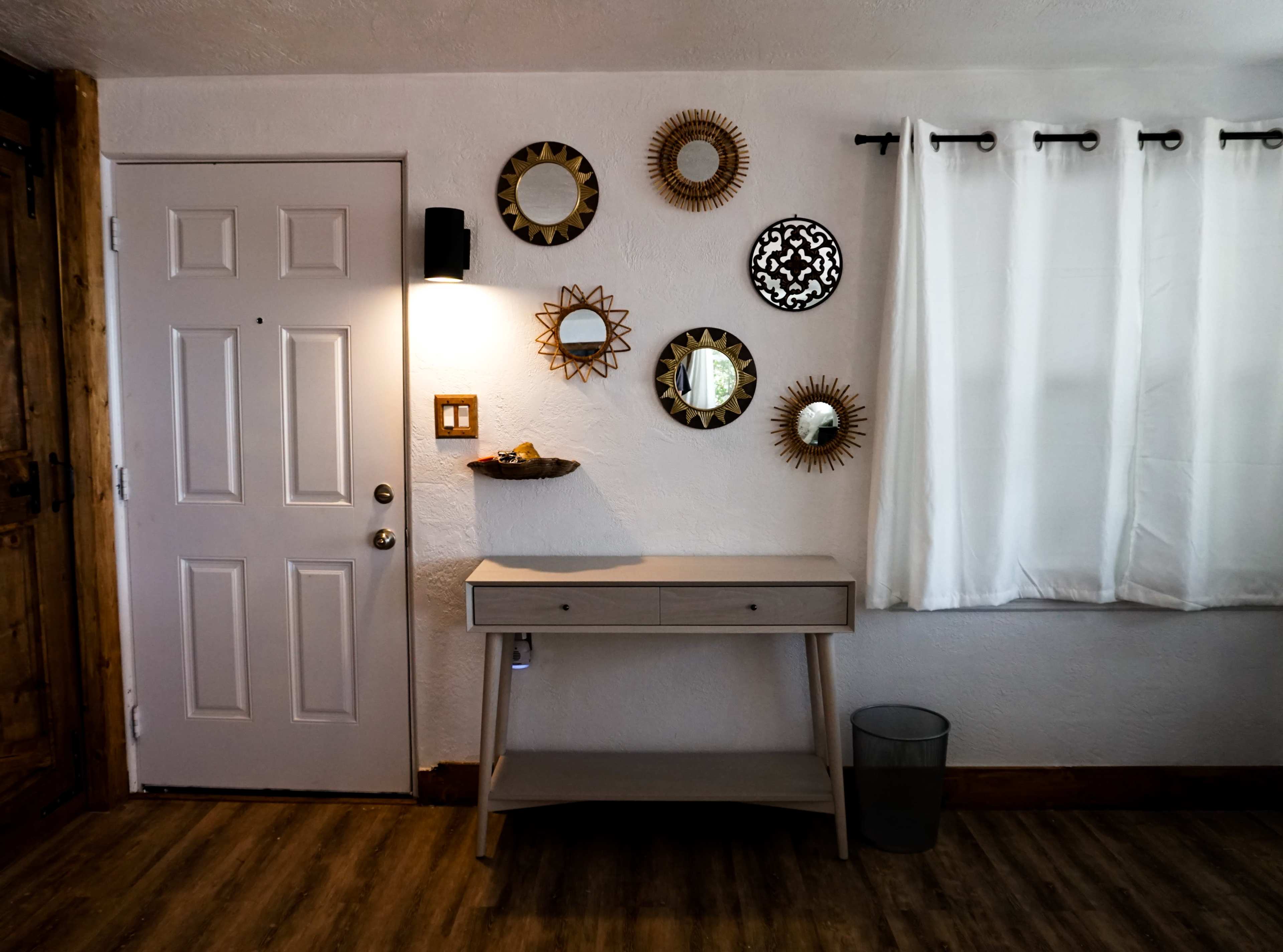 A light-colored wooden table is positioned against a wall featuring decorative mirrors and a door on the left, with a white curtain partially covering a window.
