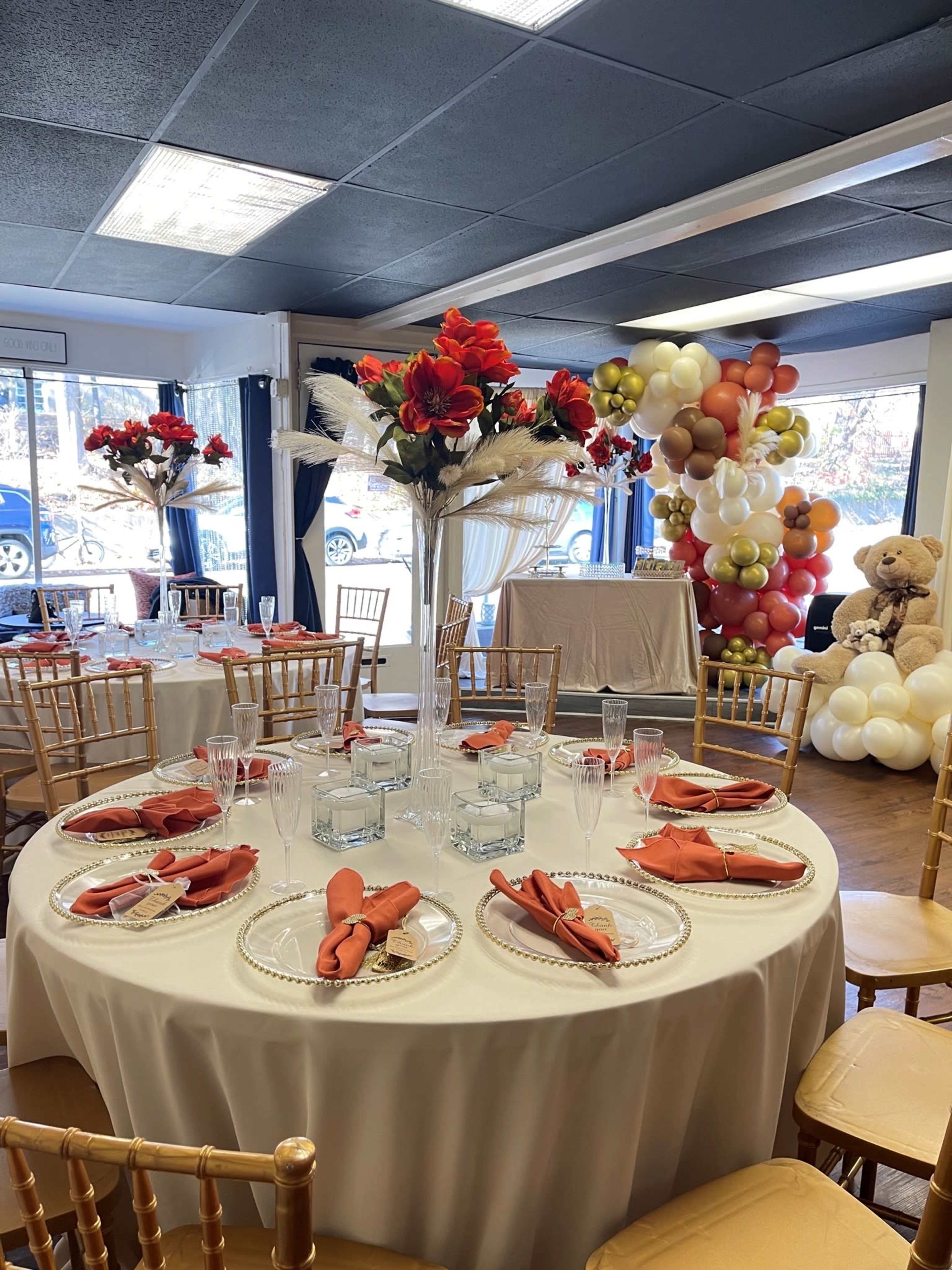 A banquet room is set with elegantly arranged tables featuring floral centerpieces, dessert plates, and decorative balloons in the background.